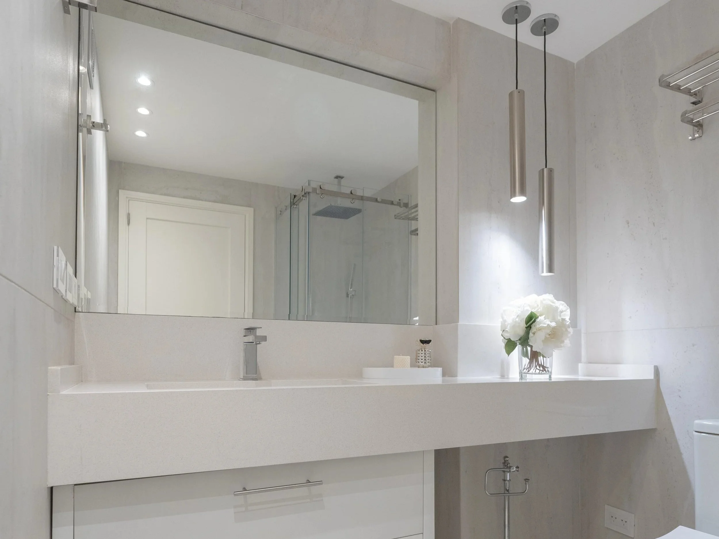 Lavabo blanco con flores blancas en un jarrón y accesorios decorativos, reflejado en un espejo grande en un baño moderno con paredes claras y lámparas colgantes de metal.