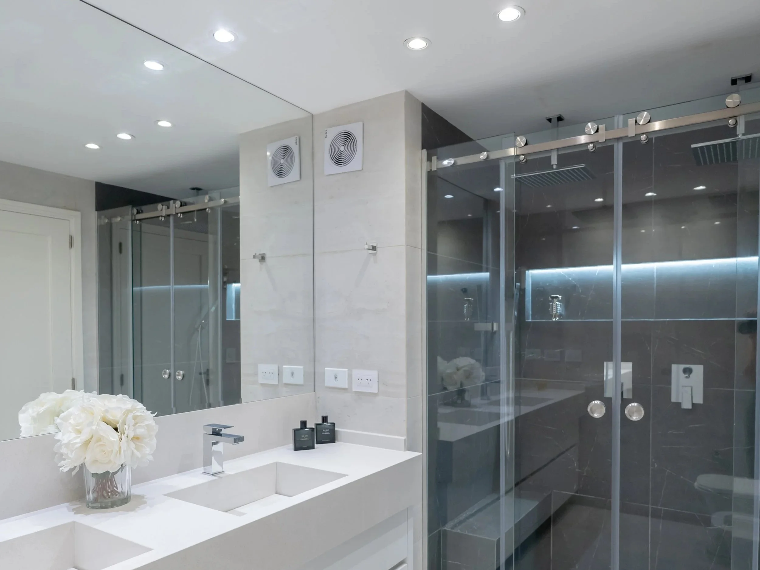 Baño moderno con lavabo de encimera blanca, espejo grande, ducha de vidrio con paredes oscuras, ventiladores en la pared, y flores blancas en un jarrón.
