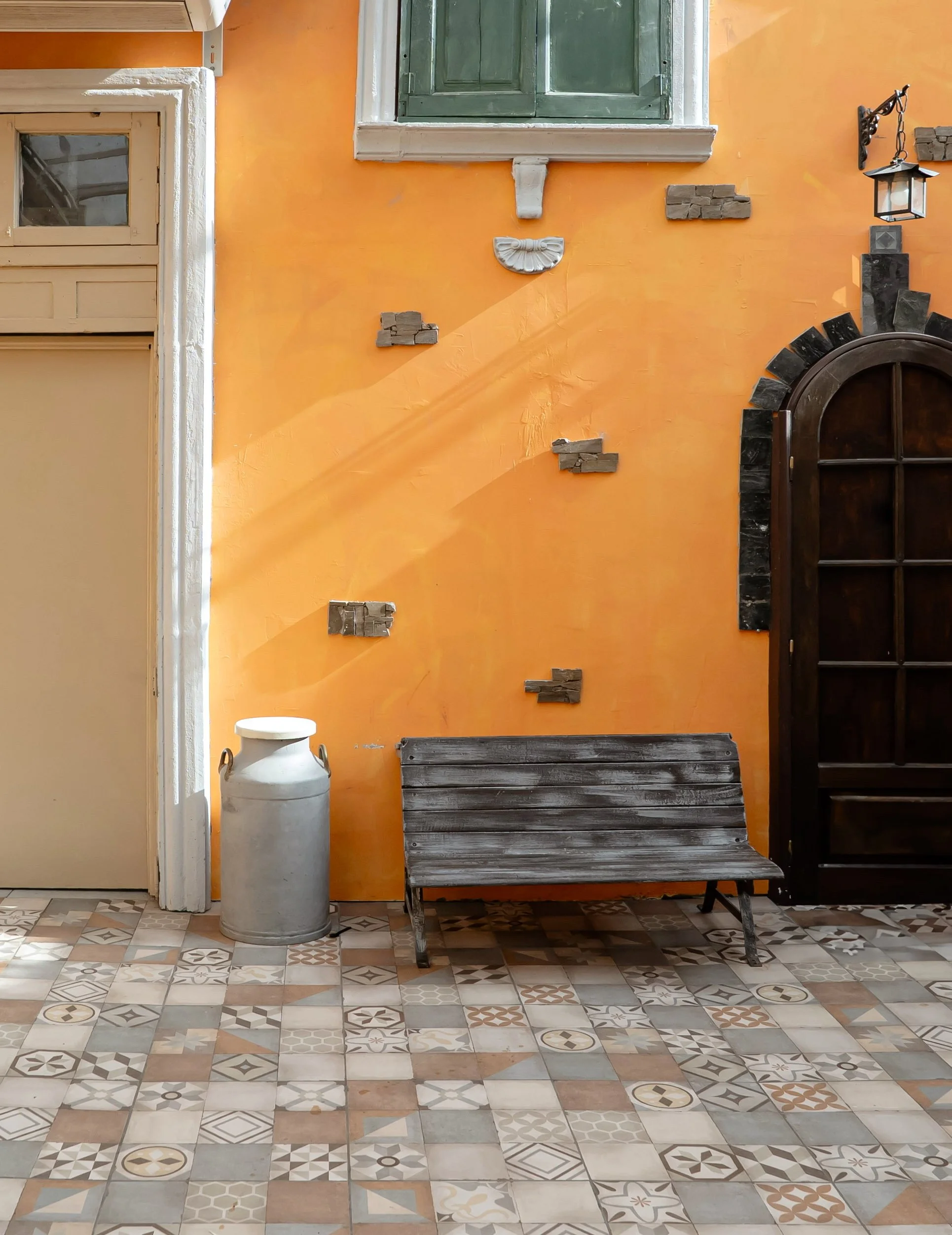 Pared naranja con ventana y detalles en colores oscuros, banca de madera envejecida, recipiente de metal, piso con azulejos decorativos, lámpara de pared.