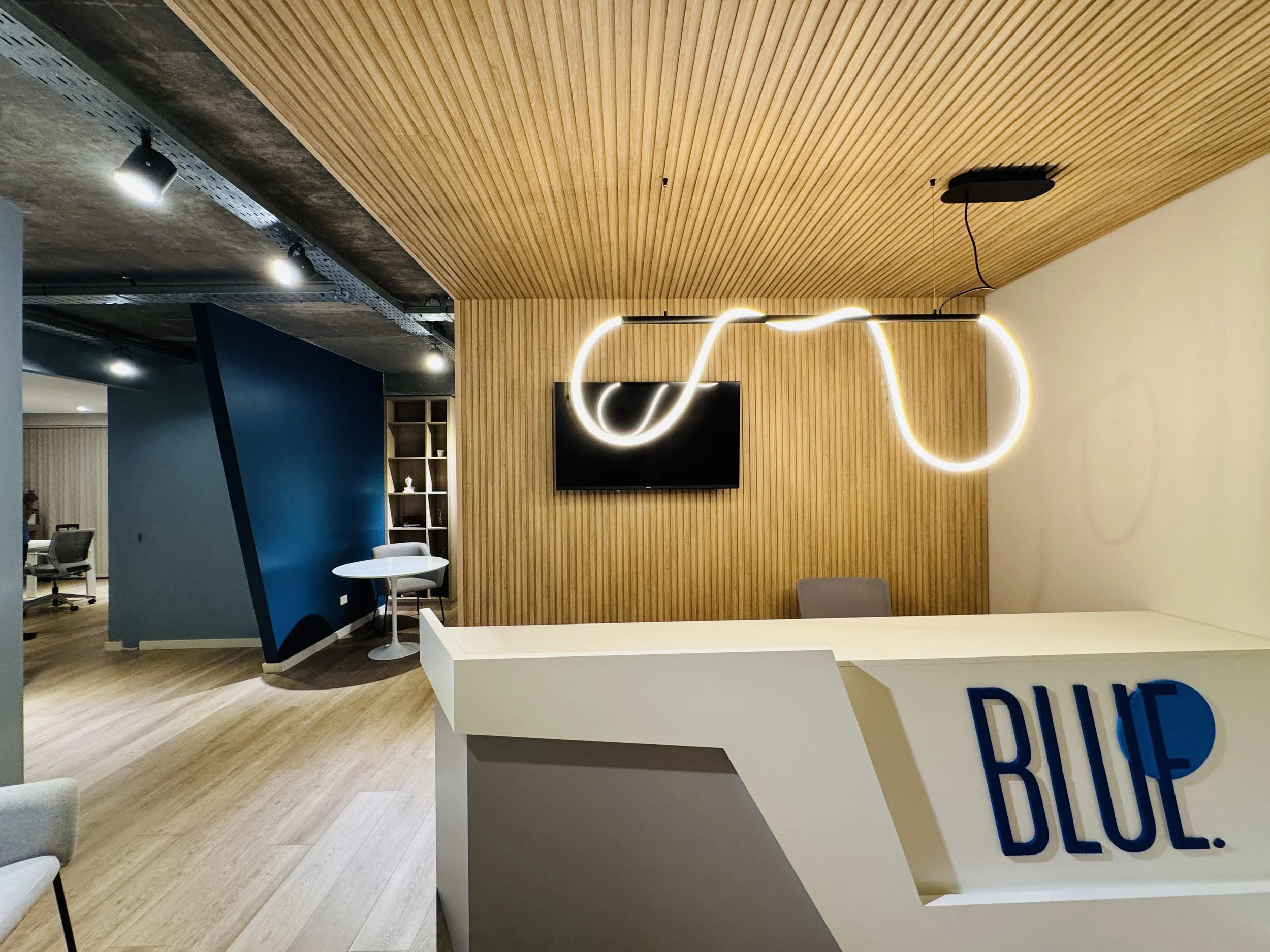 Recepción de oficina con barra blanca, cartel que dice 'BLUE.', pared de madera, pantalla en la pared, lámpara moderna colgante y área de descanso con muebles y paredes decoradas en tonos neutros y azul.