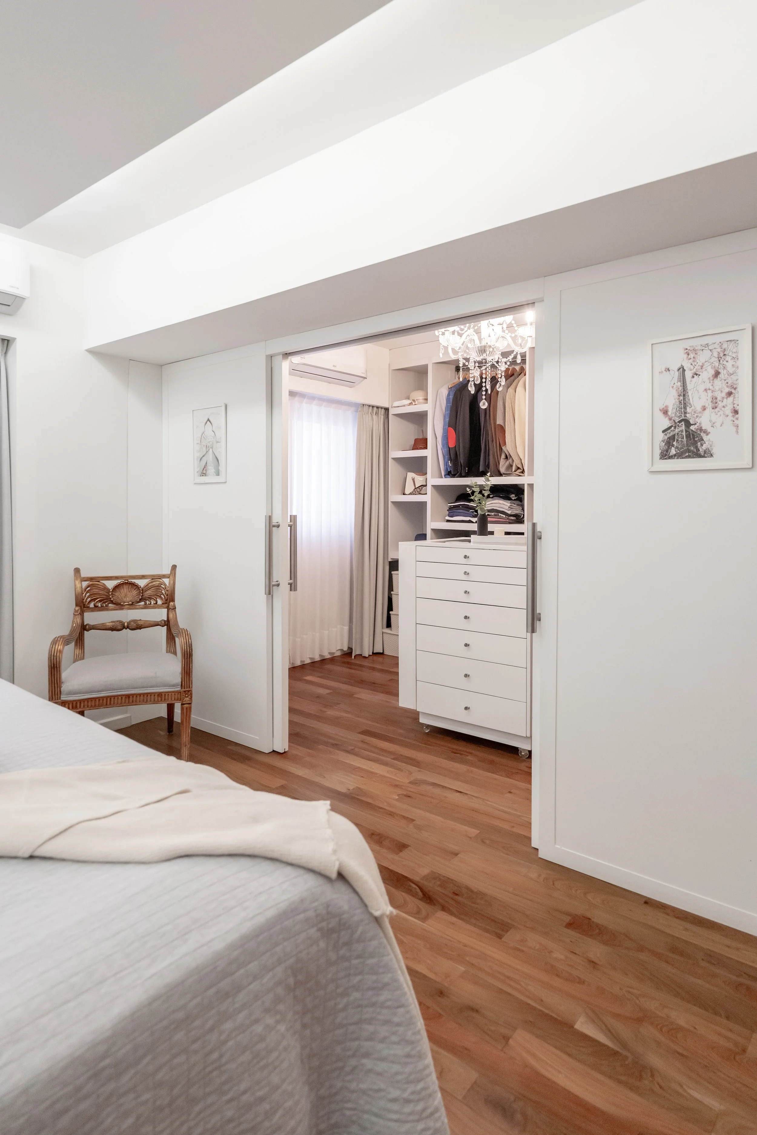 Vista de una habitación con cama, una silla de madera y un área de armario con ropa y una lámpara de araña en un espacio moderno y luminoso.