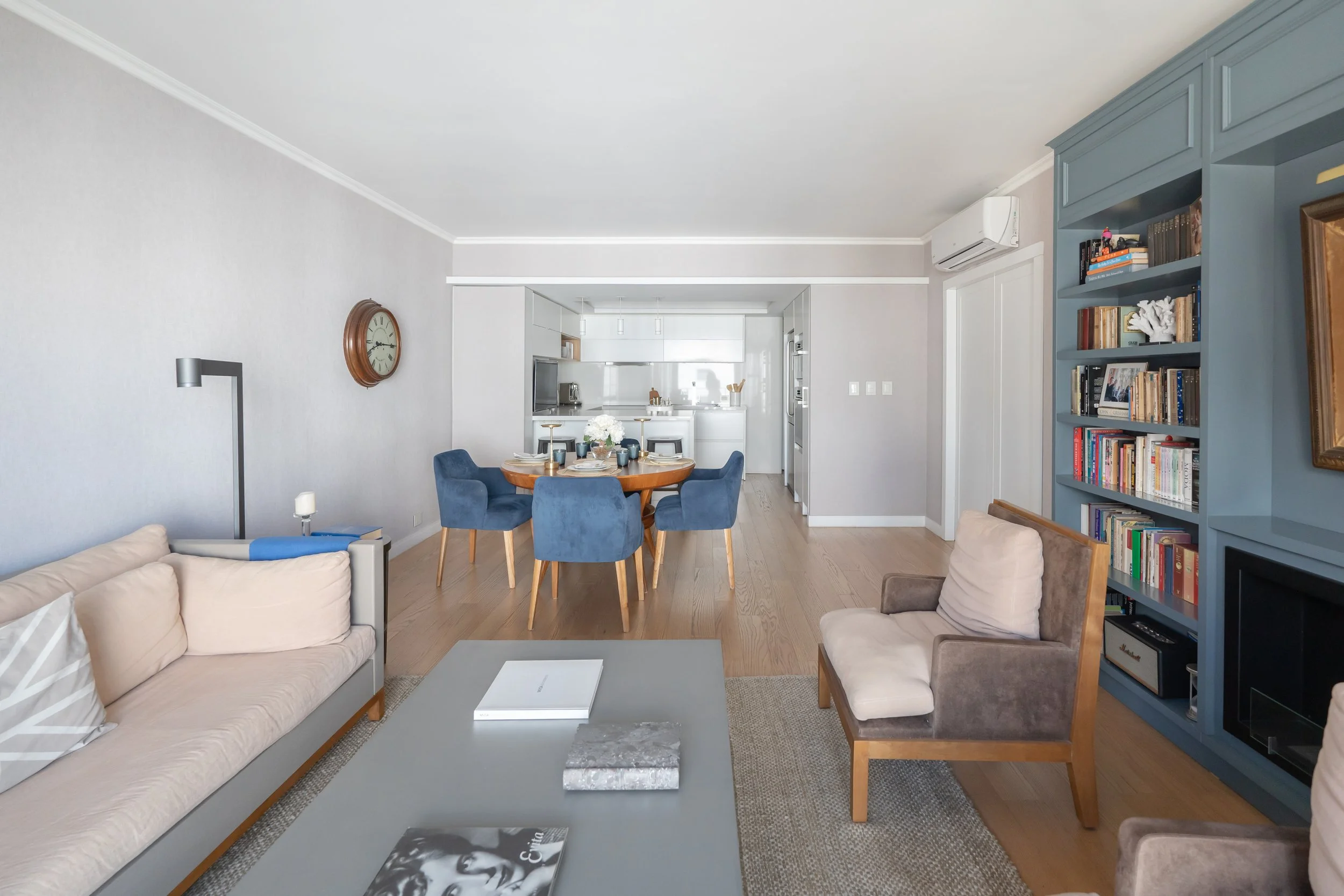 Sala de estar con sillones beige y de color marrón, librero lleno de libros, mesa de centro gris, y comedor con sillas azules alrededor de mesa de madera, cocina blanca al fondo.