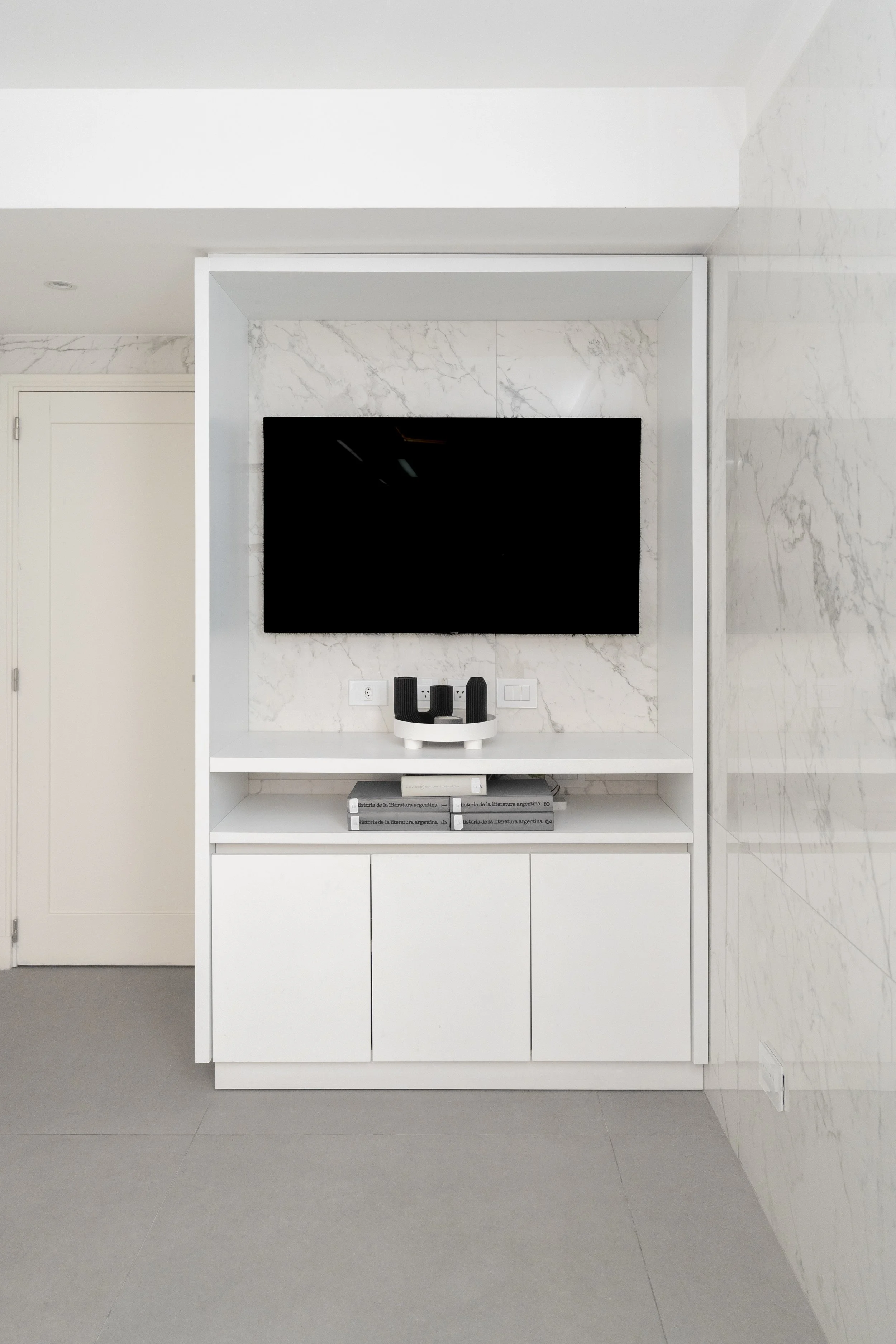 Mueble blanco con estantes, televisor negro, y algunos libros en una sala moderna con paredes de mármol.