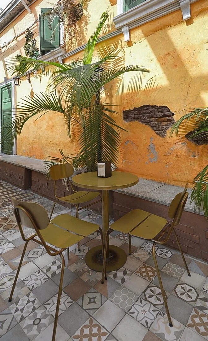 Una mesa redonda con cuatro sillas en un patio con paredes amarillas, plantas grandes y mosaicos en el suelo.