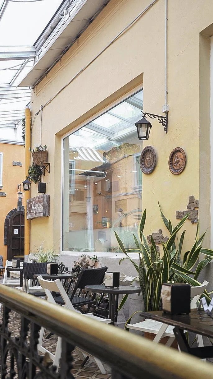 Área de restaurante al aire libre con mesas, sillas y plantas ornamentales, paredes amarillas decoradas con ventanas, faroles y arte en cerámica.