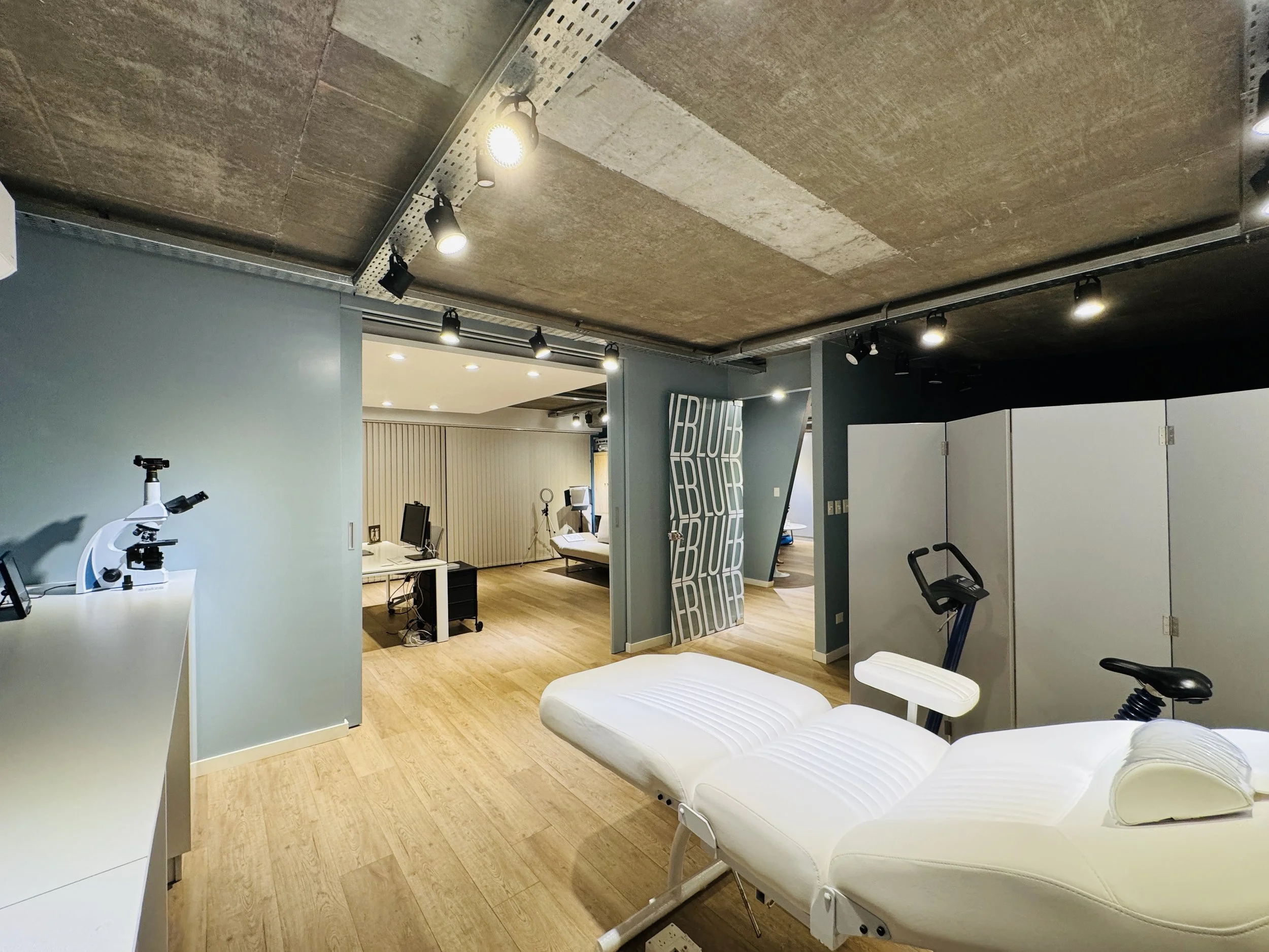 Sala de consulta médica con cama blanca, equipo de microscopio, escritorios con computadoras, y división en la zona de atención.