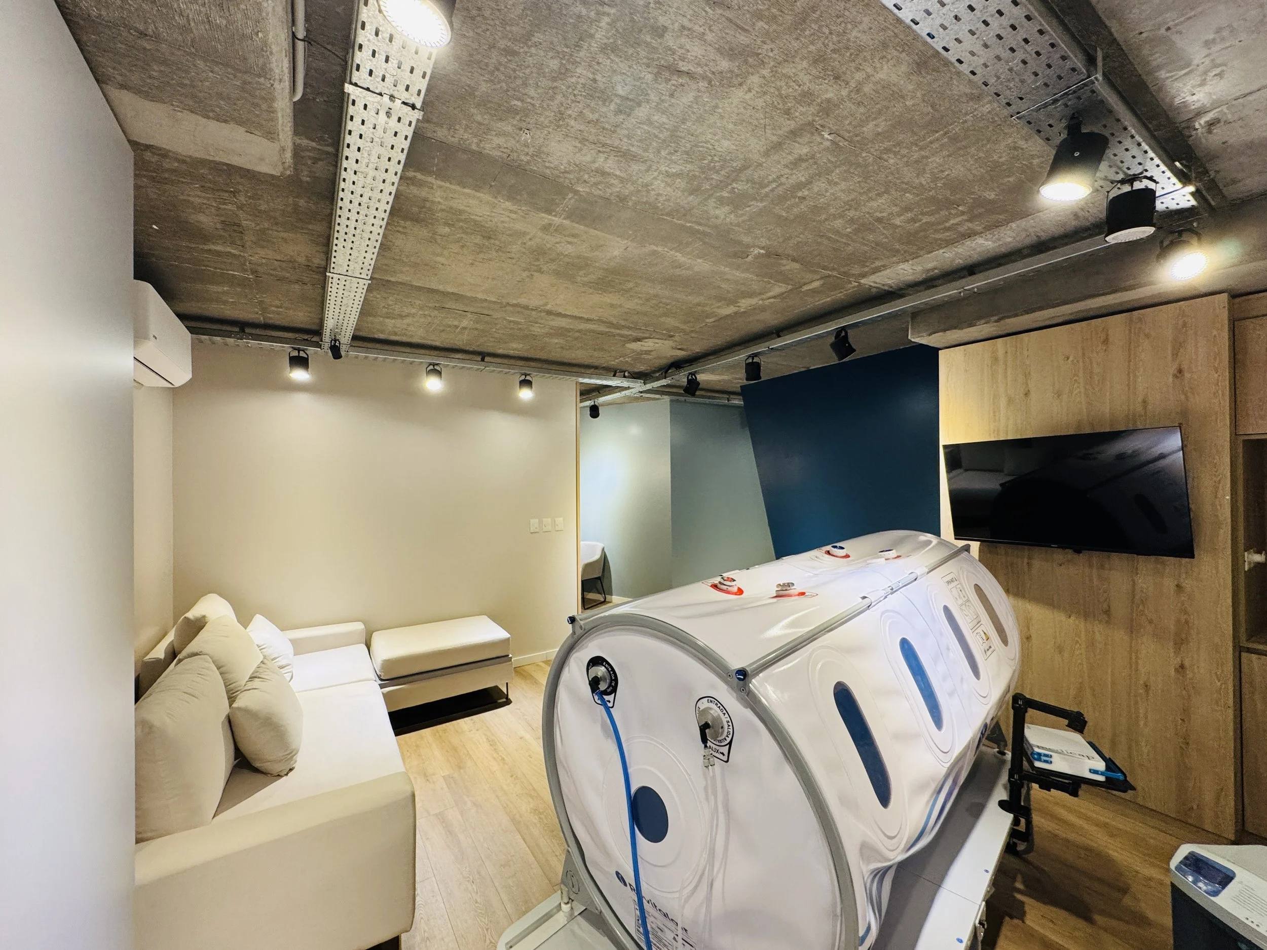 Sala moderna con sillones beige, televisión en pared de madera, equipo de crioterapia y decoración minimalista.