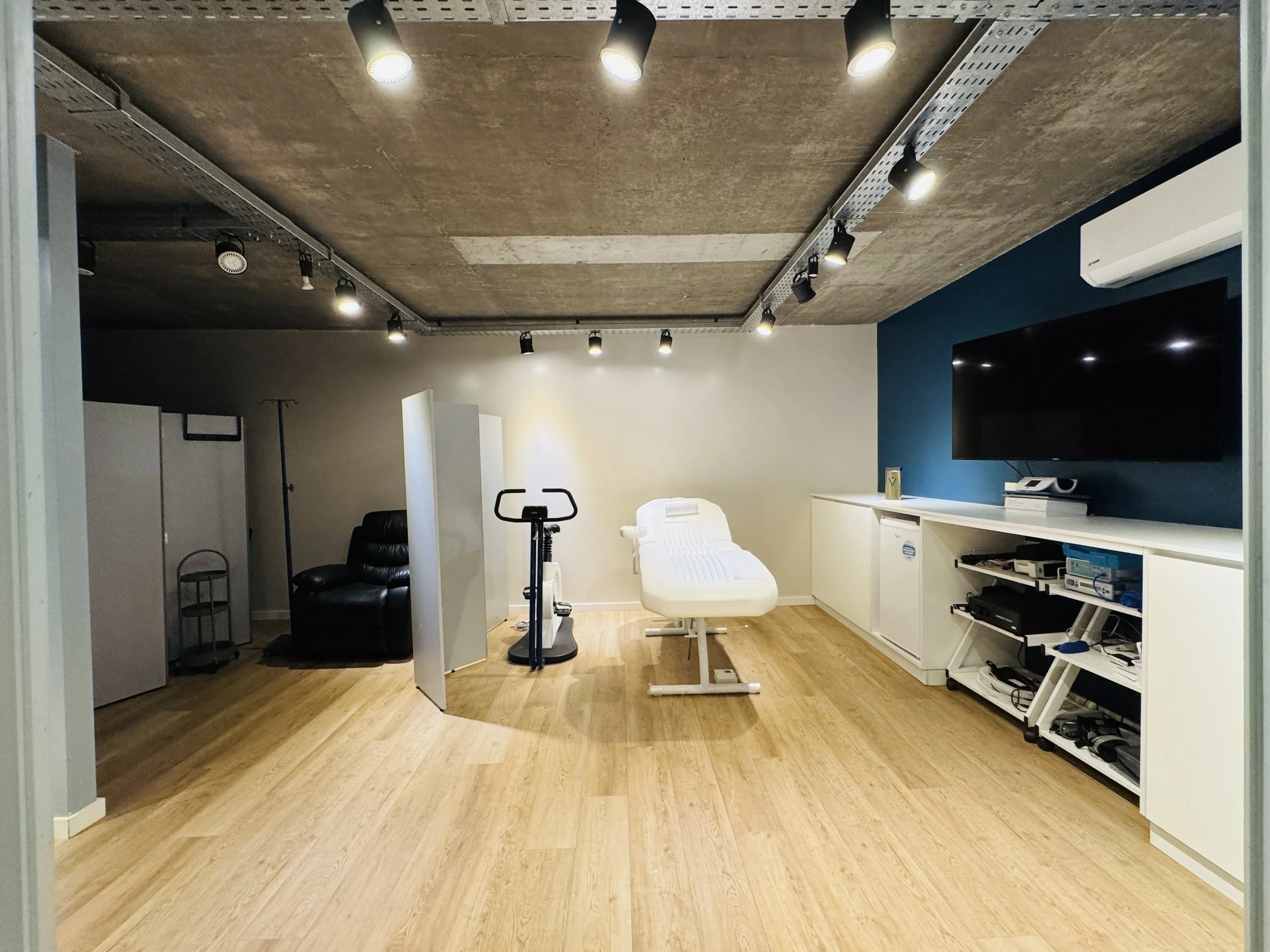 Consulta médica con cama, silla, televisor y equipo médico en una clínica moderna.