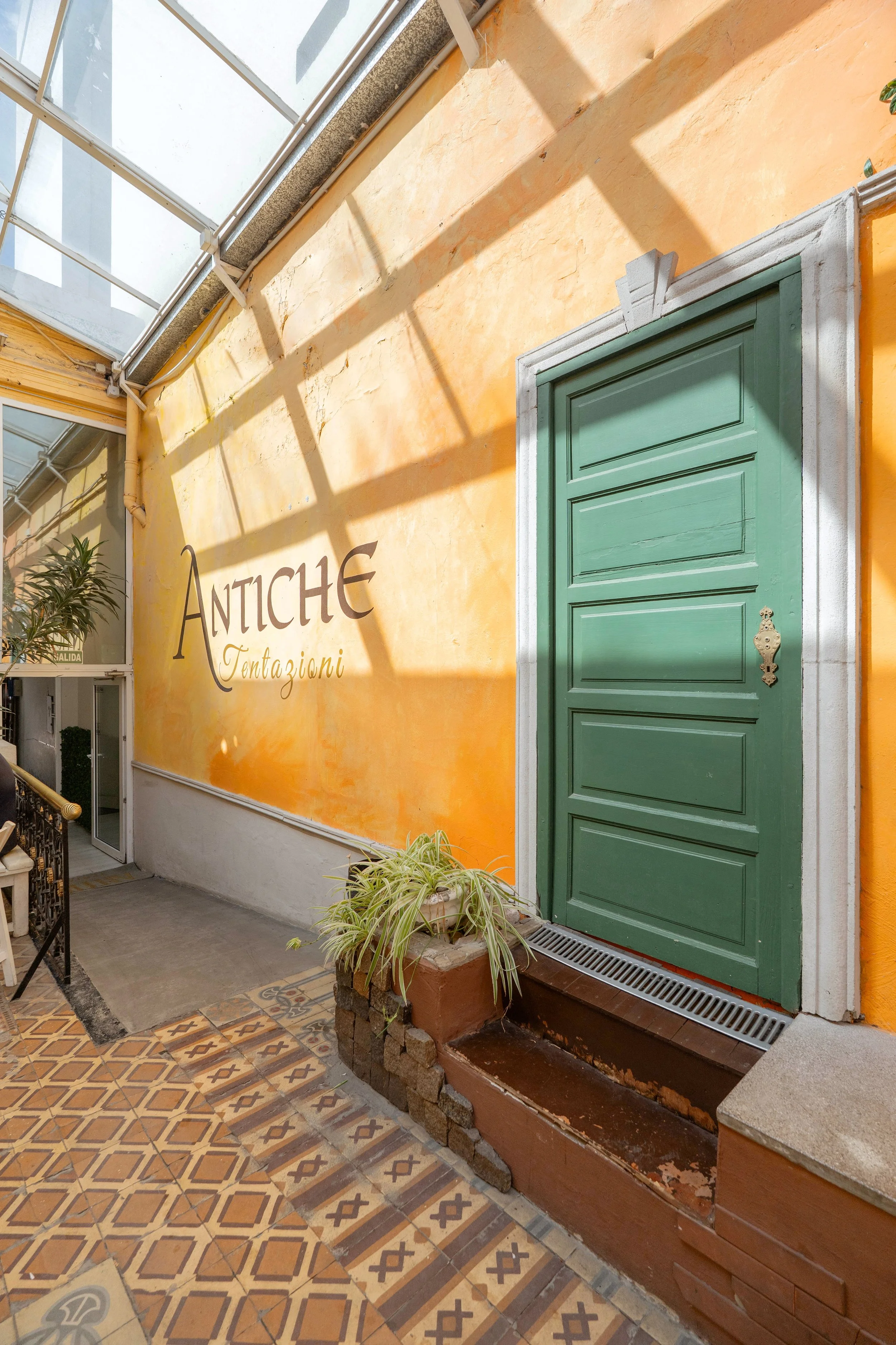 Entrada decorativa a un edificio con un muro de color amarillo, la puerta de madera verde y plantas en macetas, con letras que dicen 'Antiche Tentazioni' en la pared