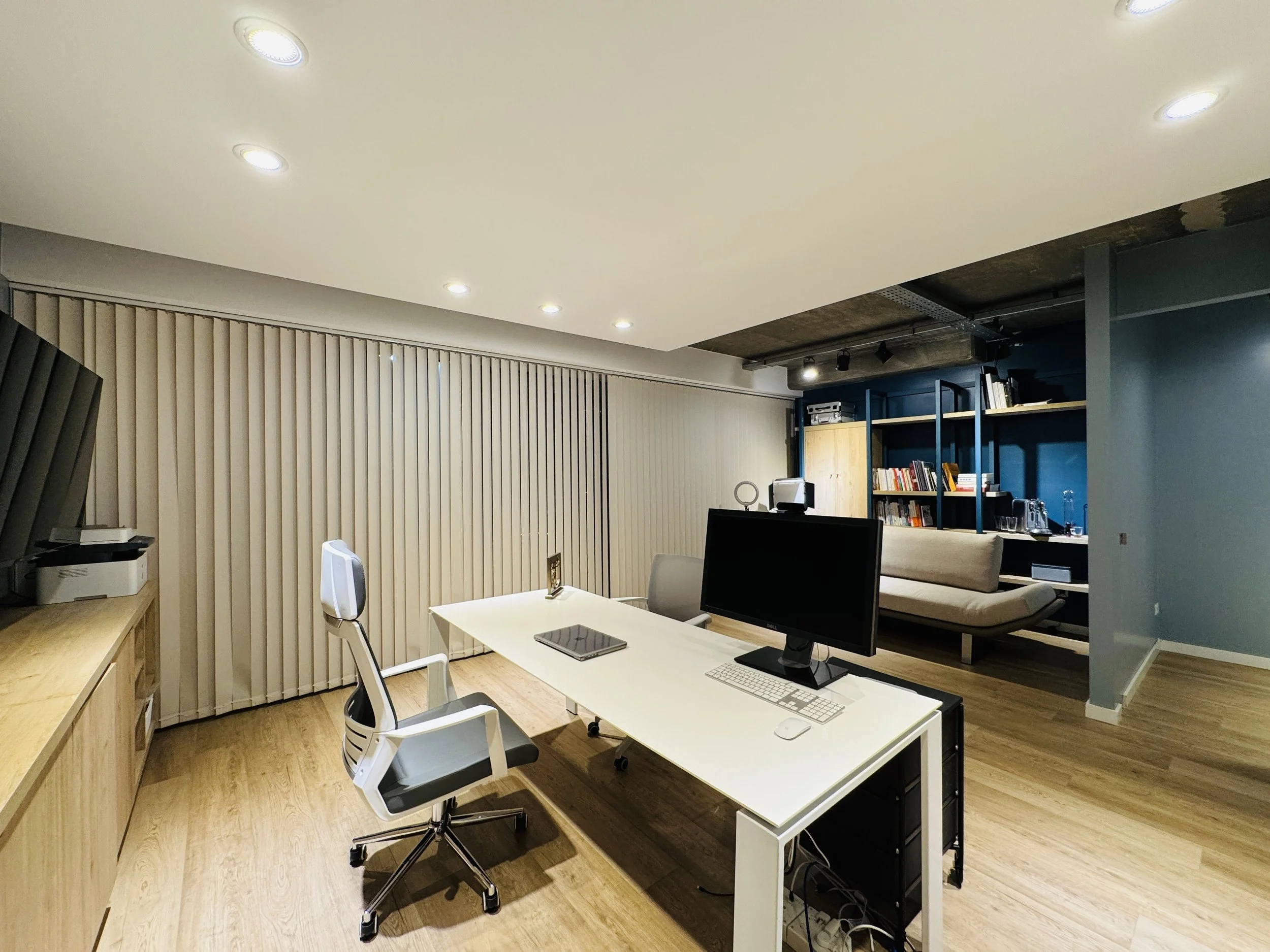 Oficina moderna con escritorio blanco, computadora, silla ergonómica, sala de lectura, libros y muebles de diseño.