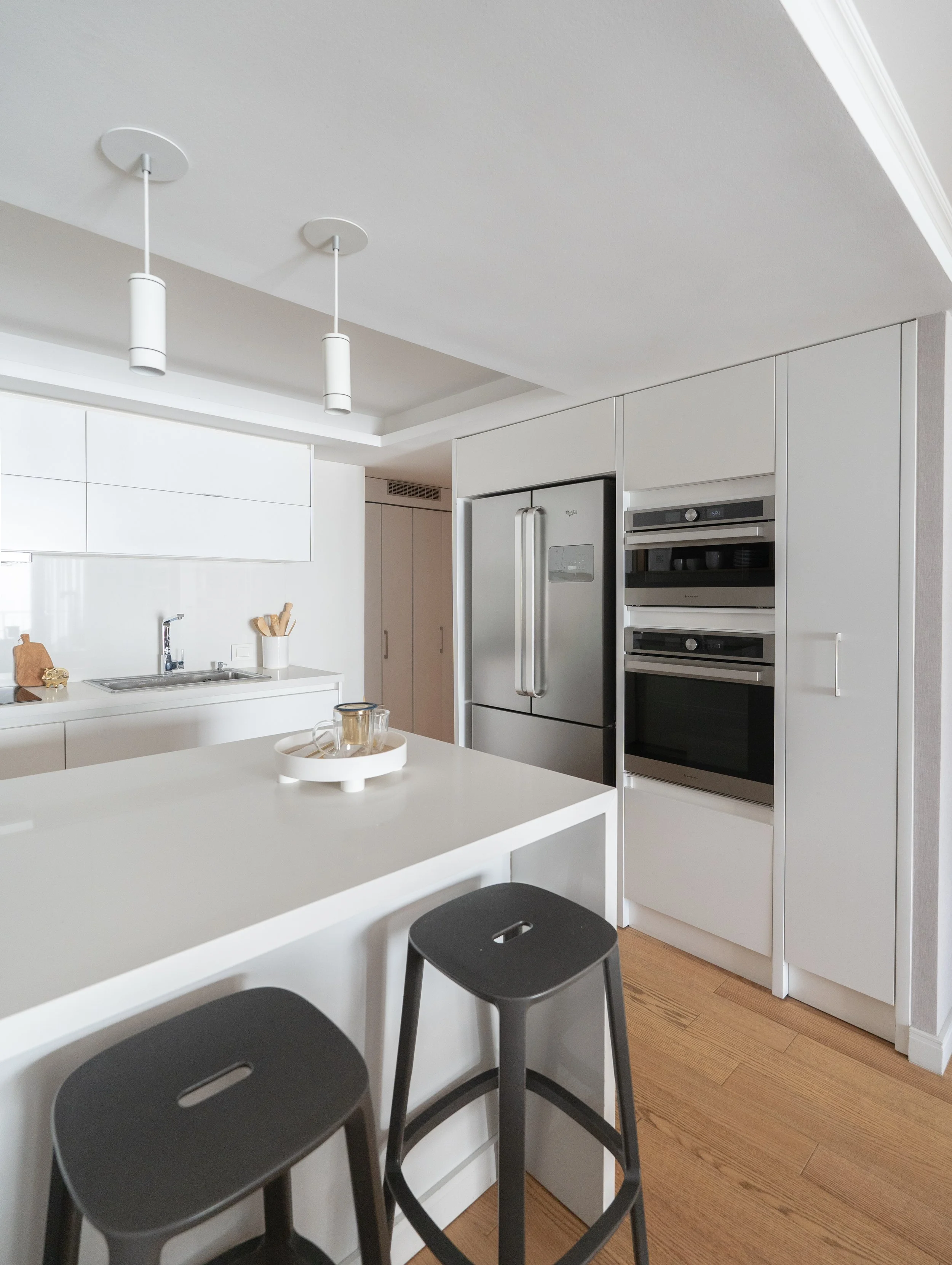 Cocina moderna con electrodomésticos de acero inoxidable y mobiliario blanco, isla central con dos taburetes negros.