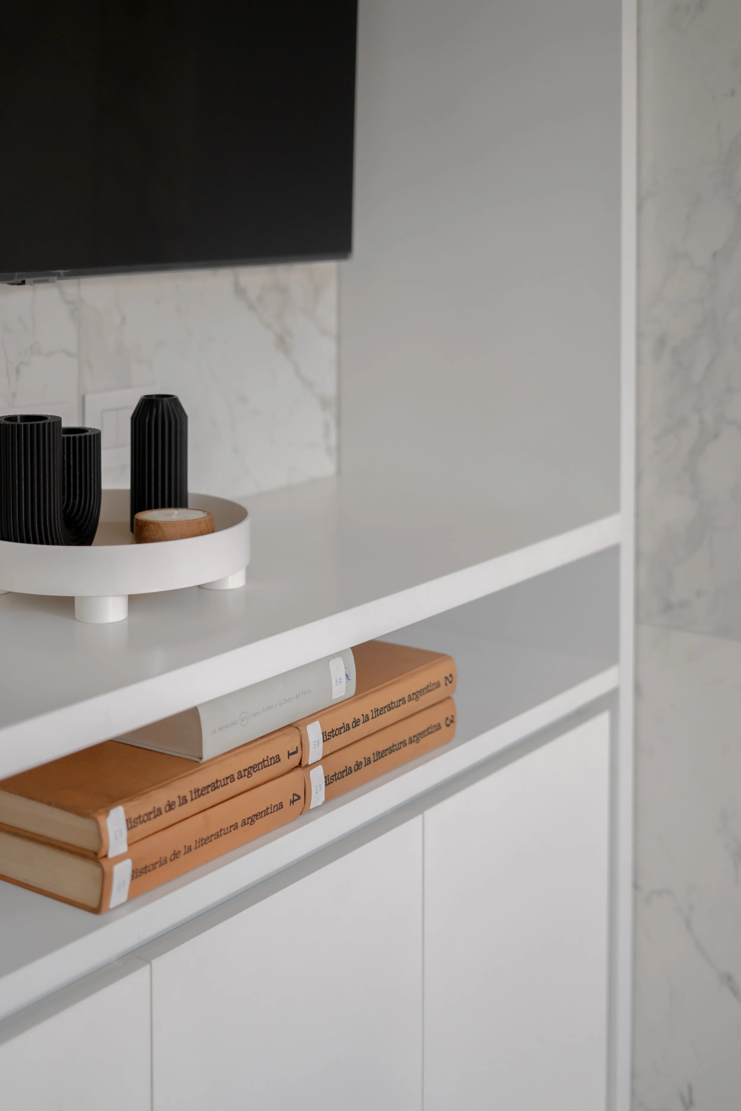 Estantería blanca con libros de historia de la literatura argentina y accesorios decorativos en la parte superior.