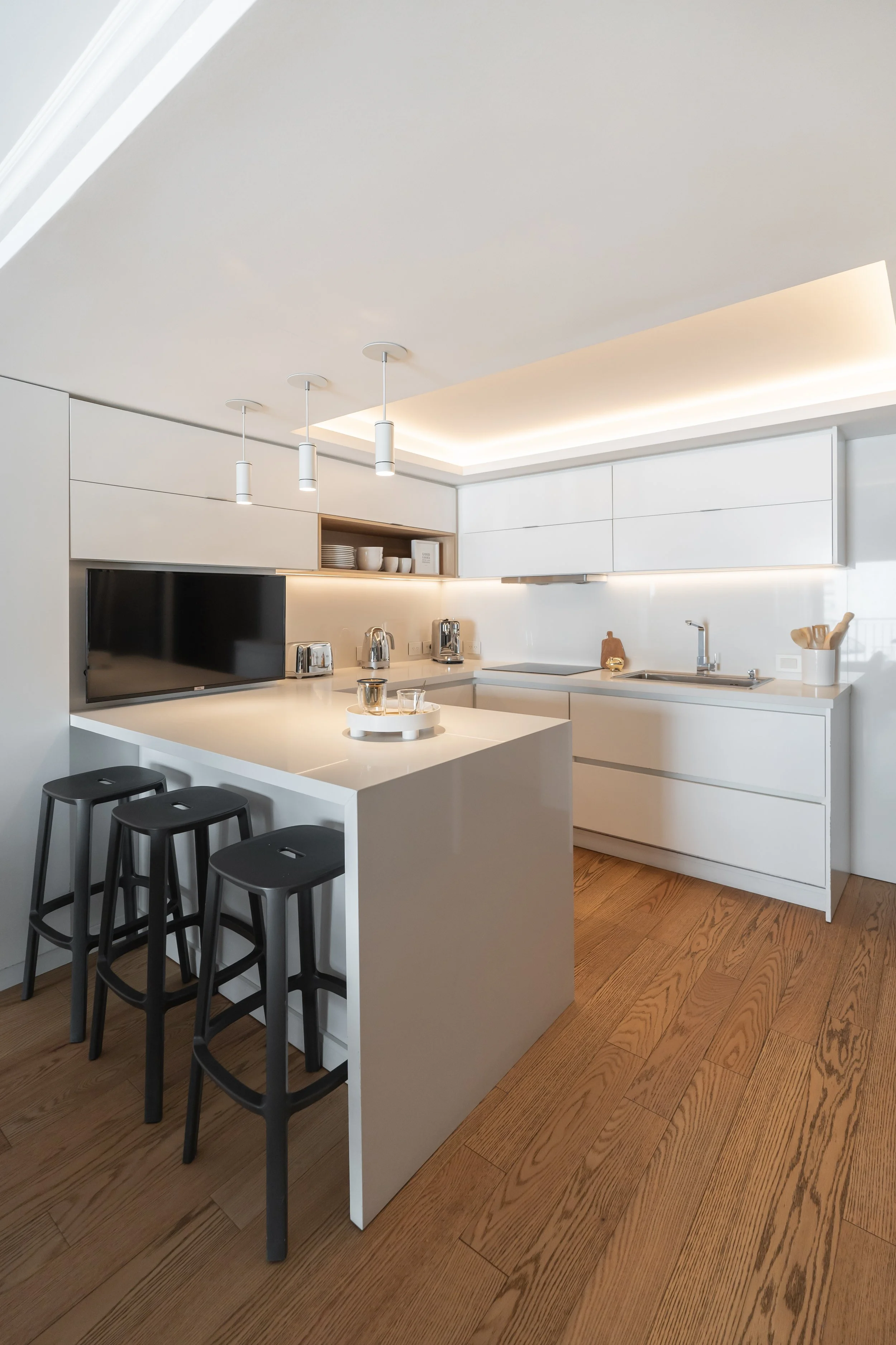 Cocina moderna, predominantemente blanca, con encimera y gabinetes blancos, tres taburetes negros en la barra, suelo de madera, y electrodomésticos en acero inoxidable, incluida una televisión.