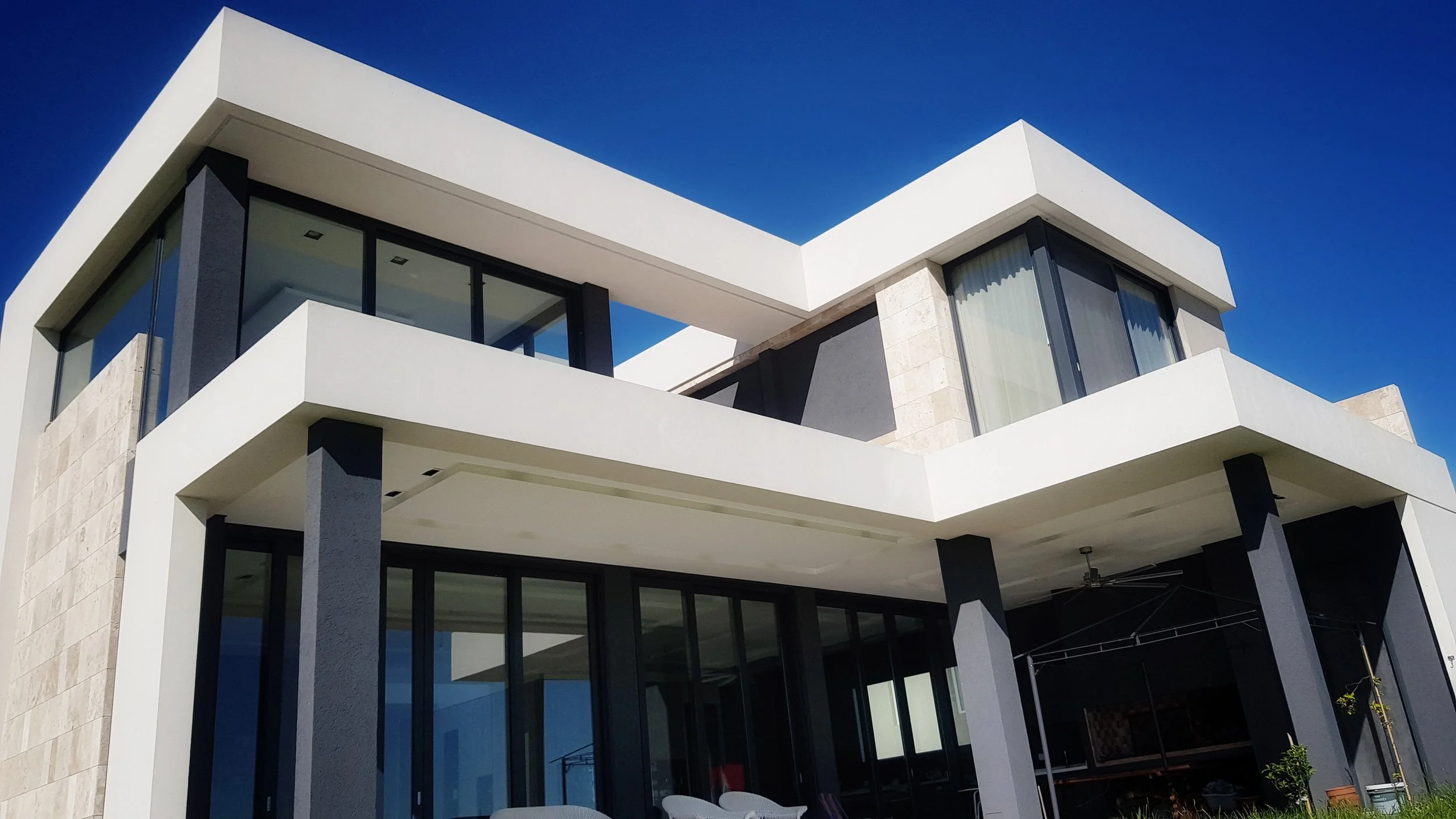 Vista moderna de una casa de varios pisos con paredes blancas, ventanas de cristal y pilares oscuros, bajo un cielo azul sin nubes.