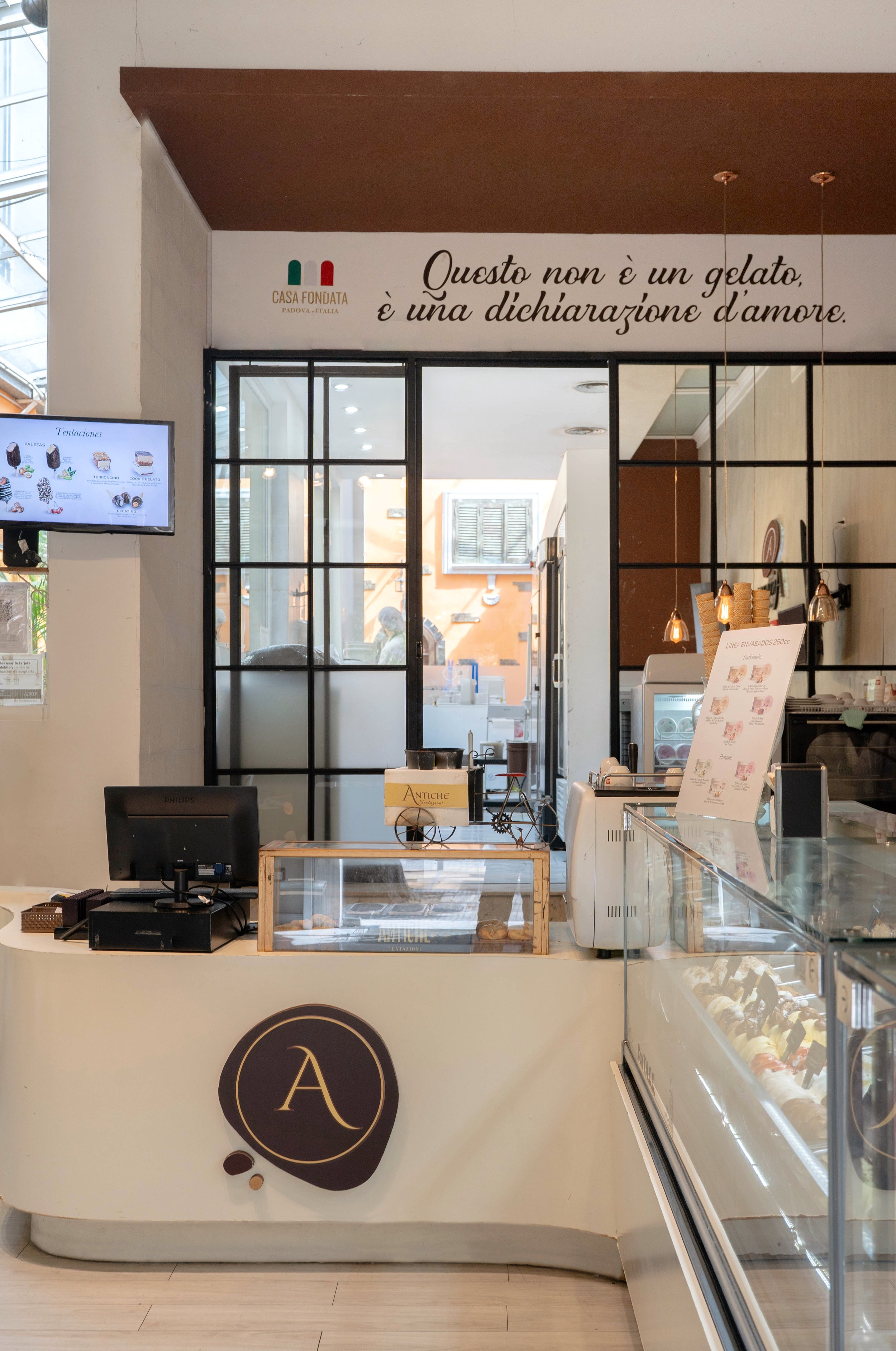 Mostrador de heladería con vitrinas de helados, caja registradora y señal de menú. En la pared, frase en italiano que dice 'Esto no es un helado, es una declaración de amor'.