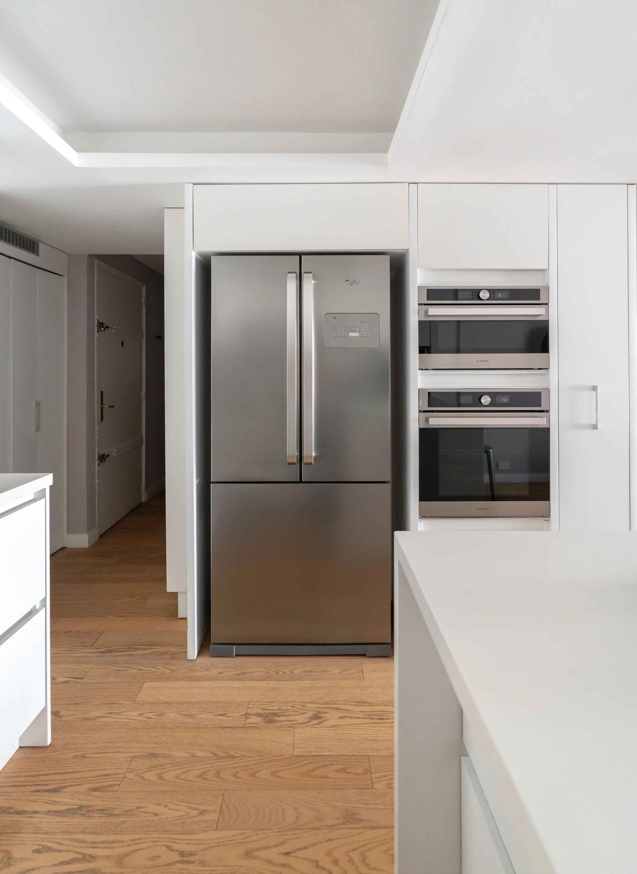 Refrigerador de acero inoxidable en una cocina moderna con armarios blancos y electrodomésticos empotrados, incluyendo hornos y microondas.