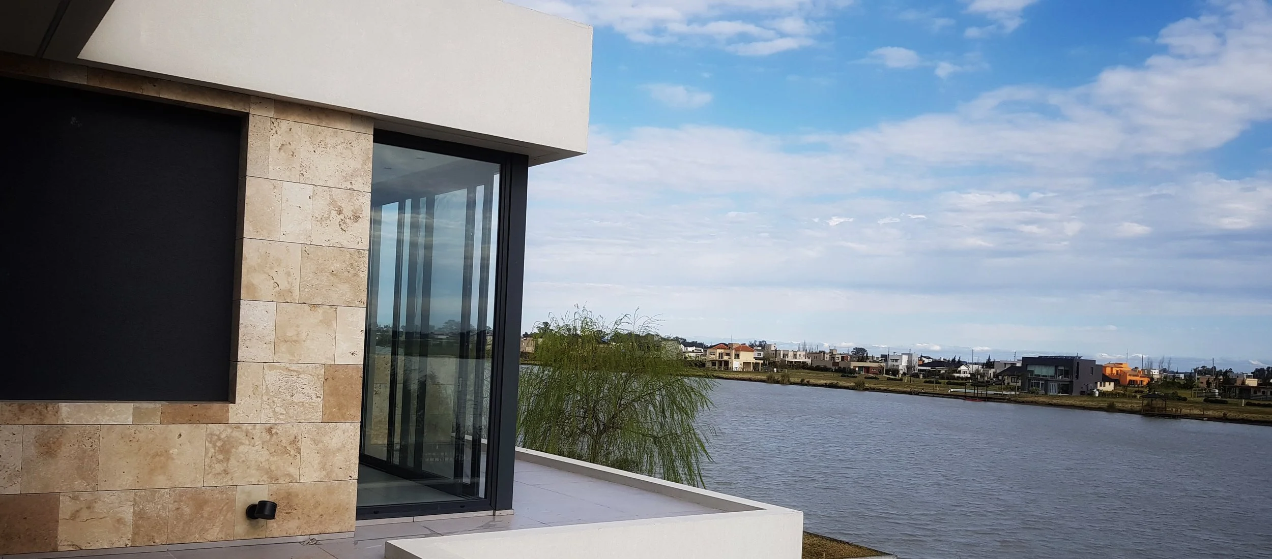 Vista de una casa moderna con paredes de piedra y ventanas grandes en un entorno junto a un río, con cielo parcialmente nublado y casas residenciales al fondo.
