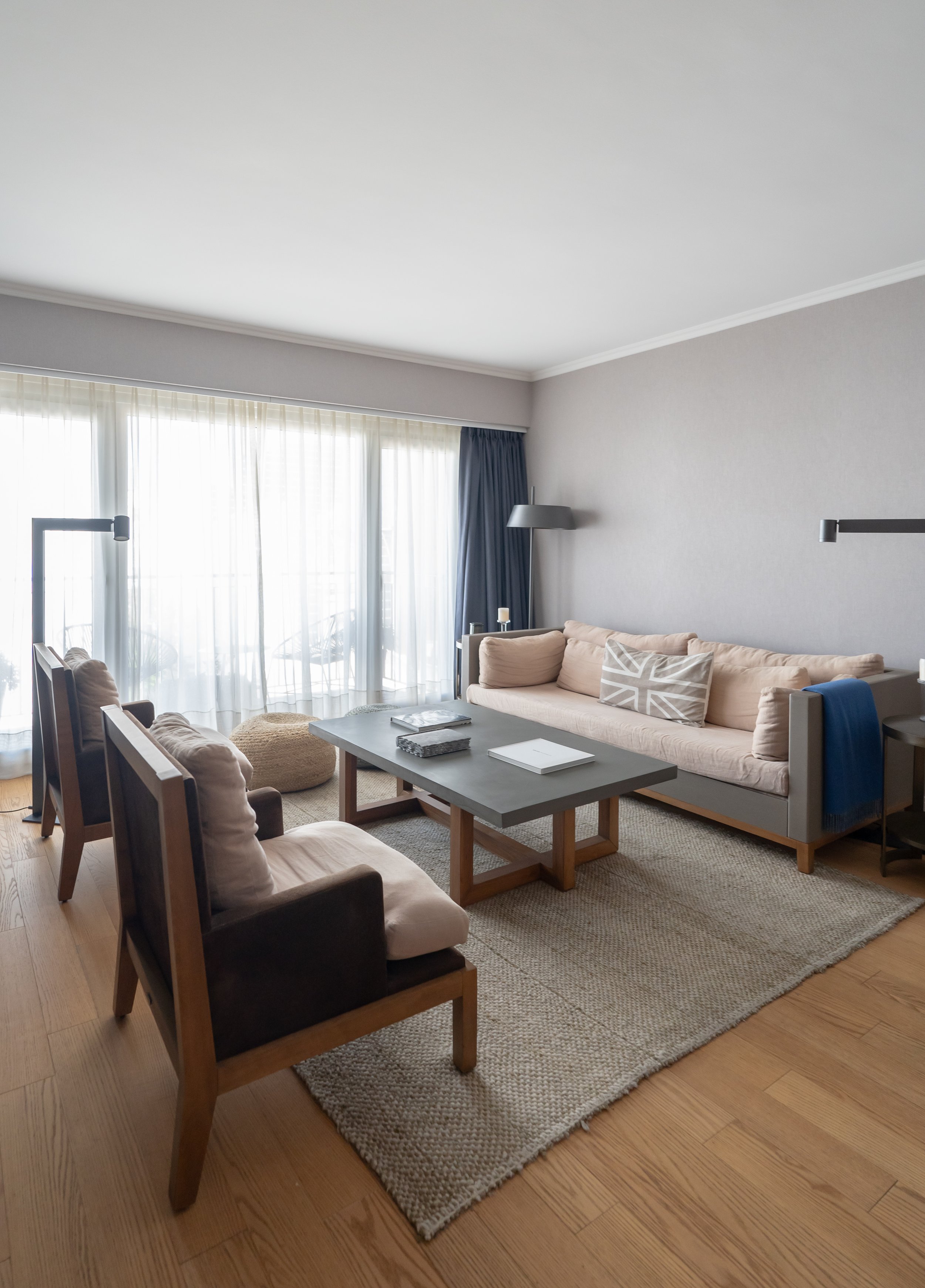 Sala de estar con sofá beige, sillas de madera, mesa de centro gris, alfombra rugosa, cortinas blancas y azules, y ventanas grandes con cortinas translúcidas.