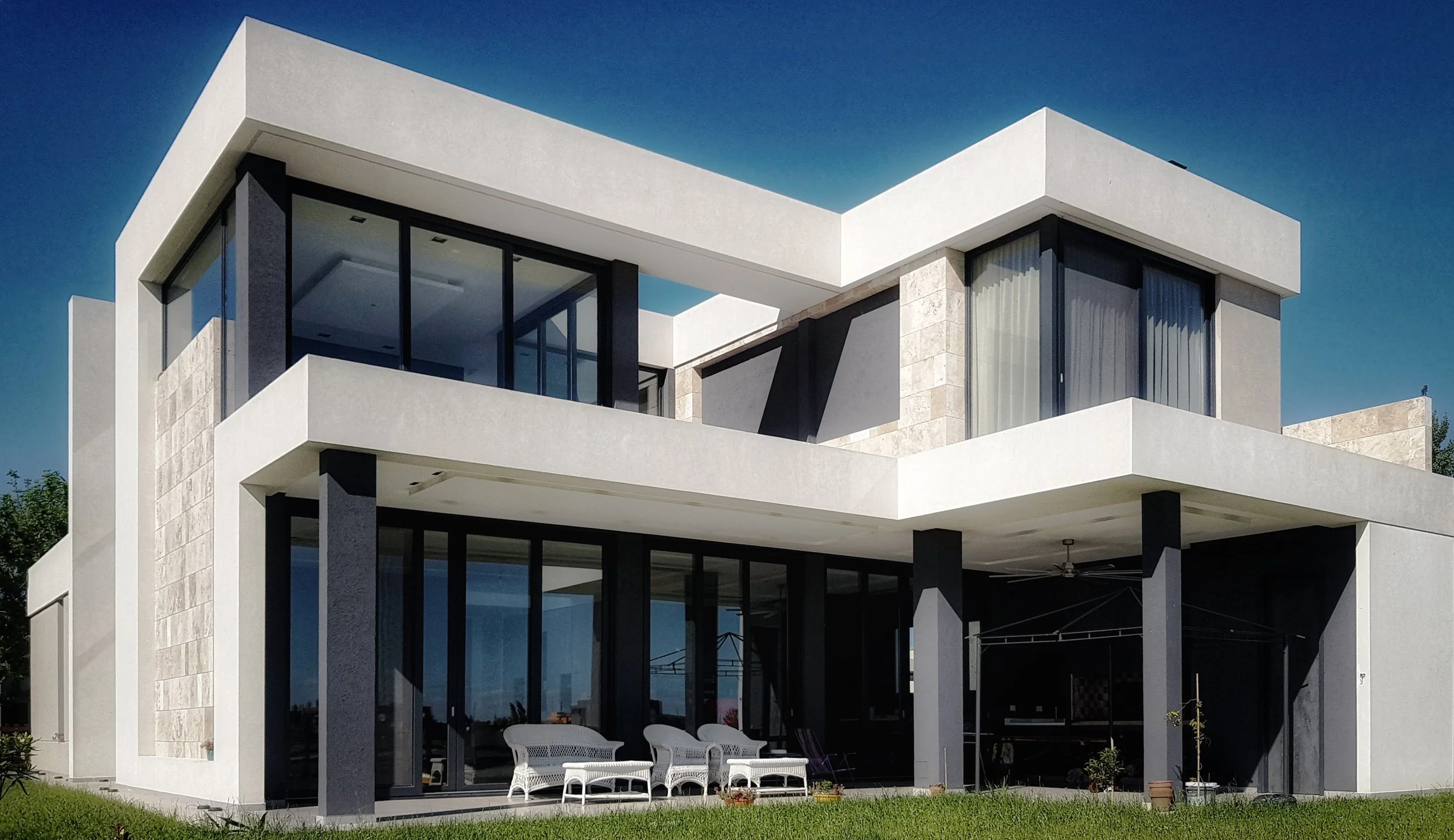 Casa moderna de dos pisos con estructura en color blanco y negro, grandes ventanas de cristal y balcón en el tercer piso, con jardín en la parte frontal.
