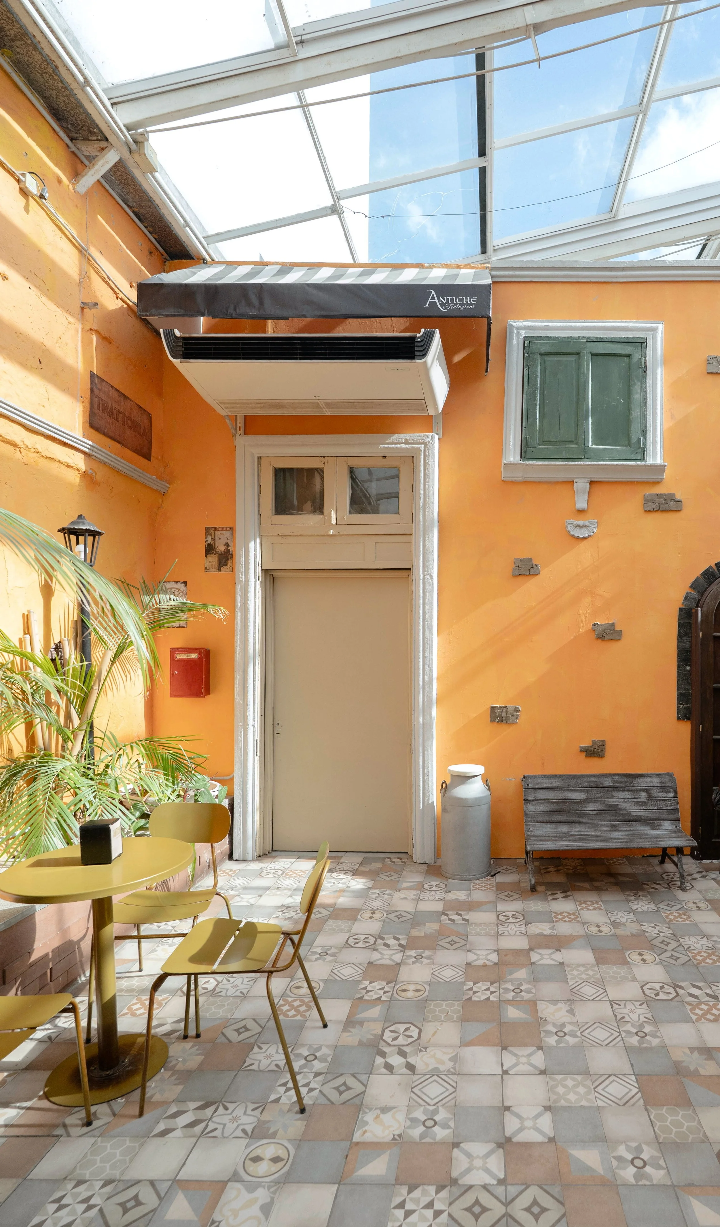 Interior de un patio con paredes naranjas, suelo de azulejos decorativos, una mesa y sillas amarillas, plantas verdes, una ventana con celosía y un banco de madera, en un ambiente soleado con estructura de vidrio en el techo.