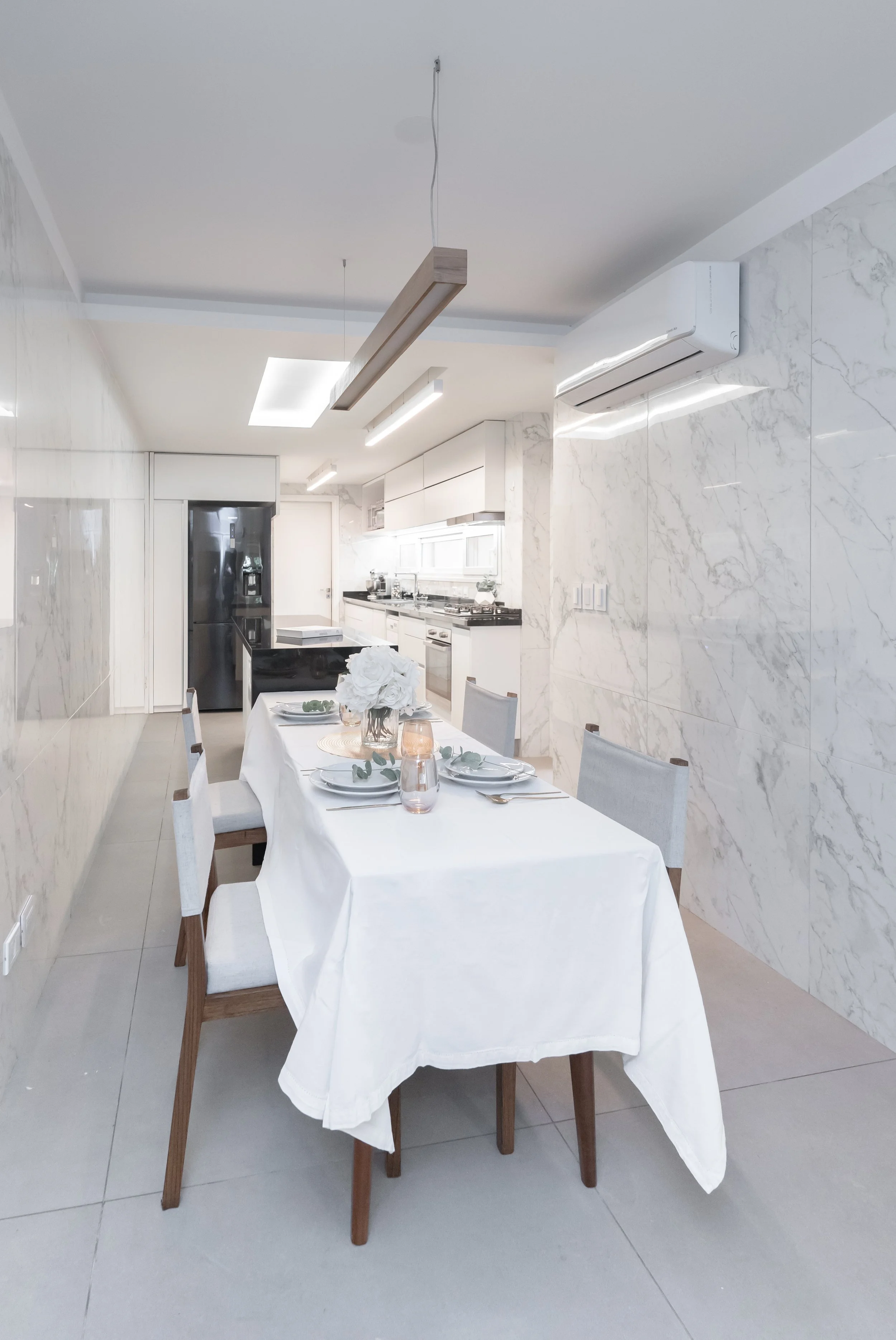 Comedor moderno con mesa cubierta con mantel blanco, decorada con flores blancas y velas, en una cocina blanca con paredes de mármol y electrodomésticos de acero inoxidable.