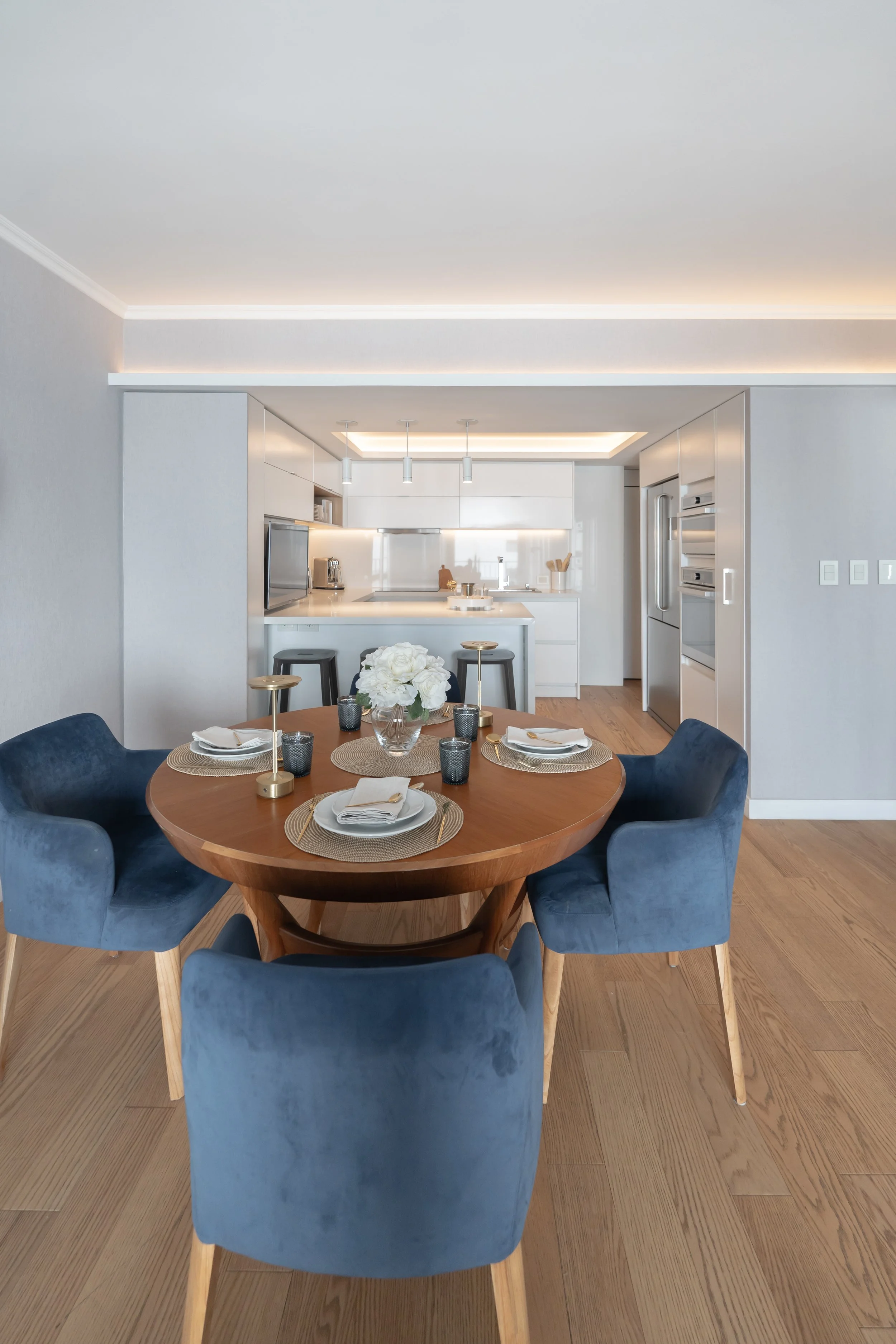 comedor con mesa redonda de madera rodeada de cuatro sillas de terciopelo azul, en el centro florero con flores blancas, platos, cubiertos y vasos, en fondo cocina moderna blanca con electrodomésticos y encimeras, suelo de madera clara