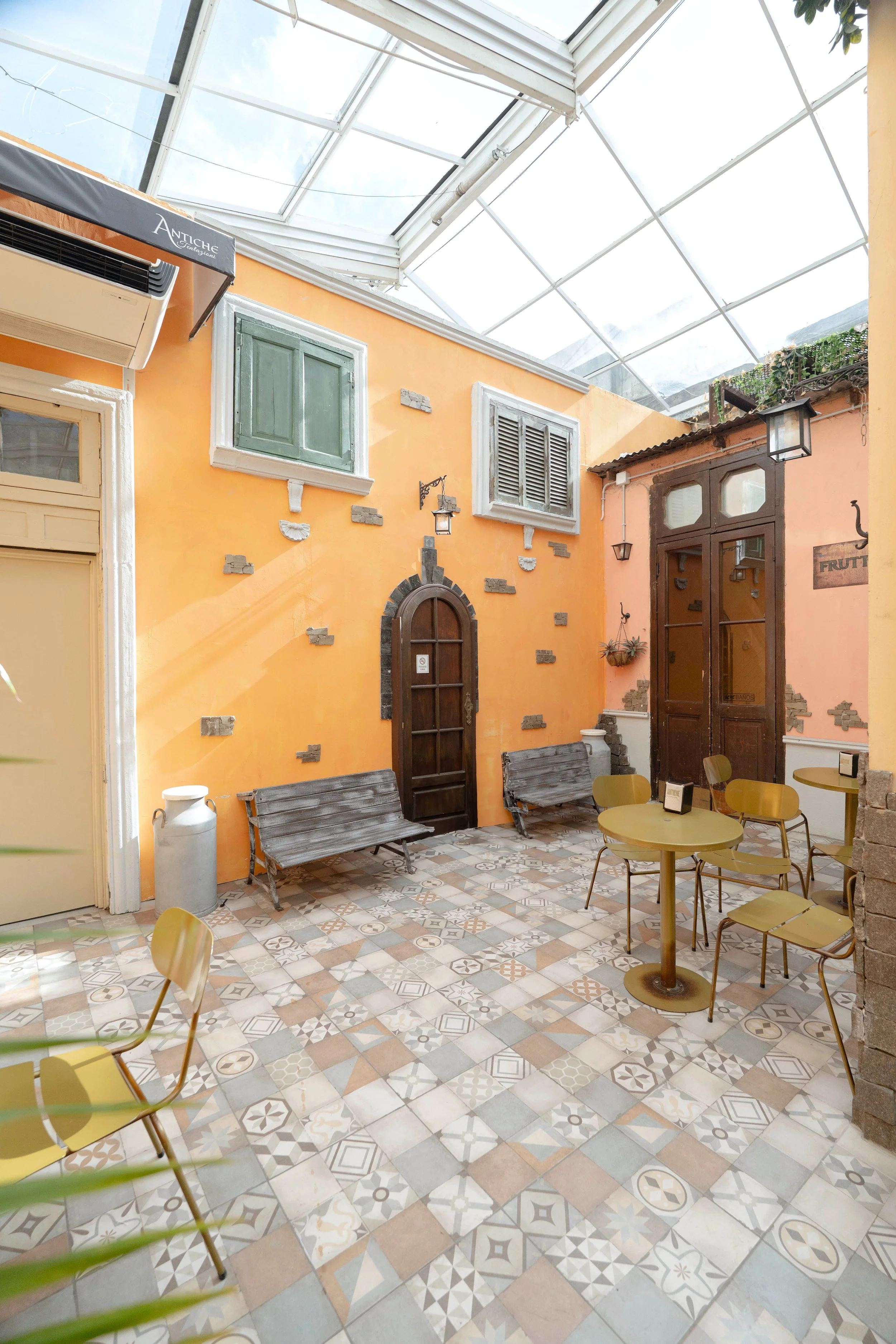 Interior de un patio decorado con paredes de colores cálidos, ventanas con marcos de madera con persianas, lámparas de pared, asientos de madera envejecida y mesas redondas de color verde oliva, con un techo de cristal que permite la entrada de luz n