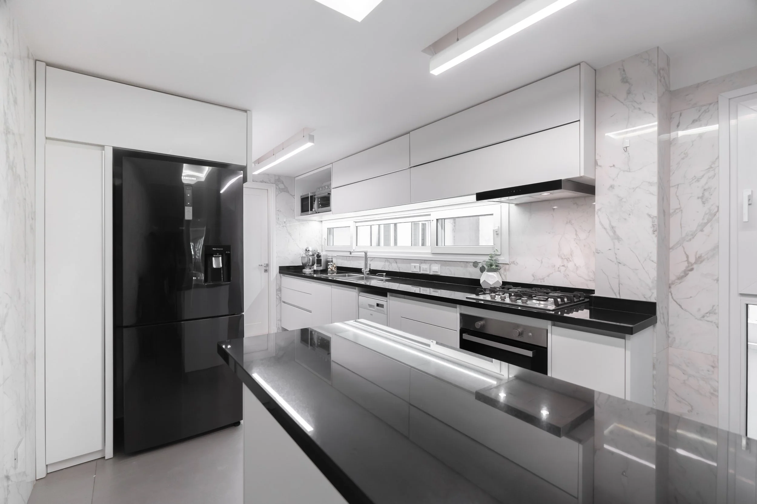 Cocina moderna con muebles blancos, encimeras negras, electrodomésticos negros, y paredes de mármol, con iluminación LED en techos y una isla en el centro.