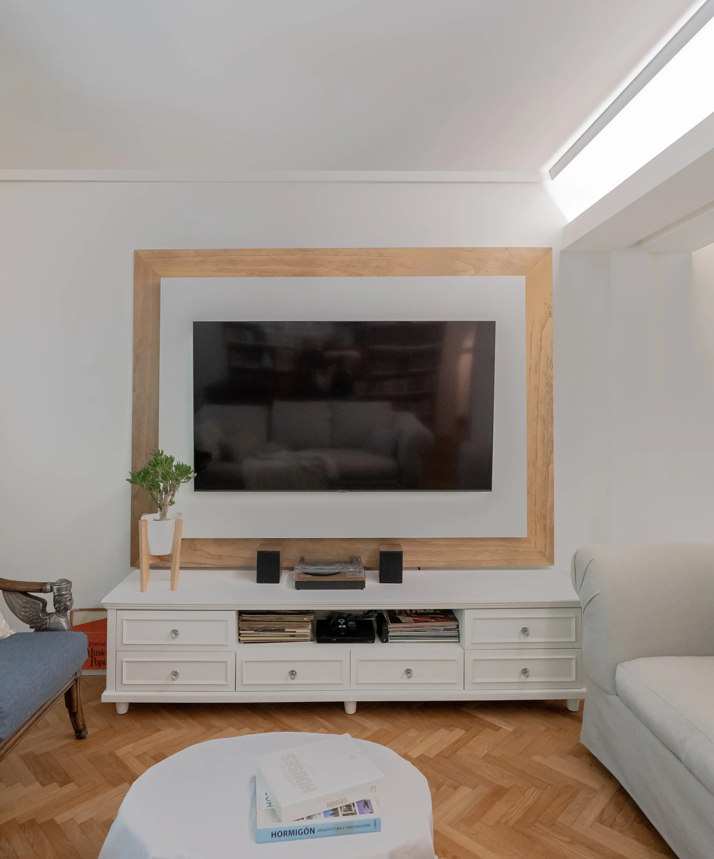 Sala de estar con televisor grande en pared, mueble blanco con libros y objetos decorativos, piso de madera, sofá blanco y silla con detalles en madera.