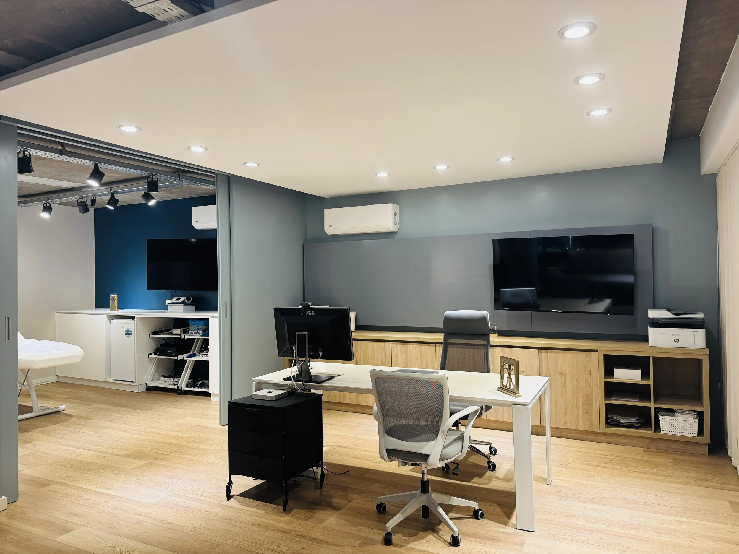 Oficina moderna con mesa, dos sillas, computador y televisión en la pared, suelo de madera y aire acondicionado.