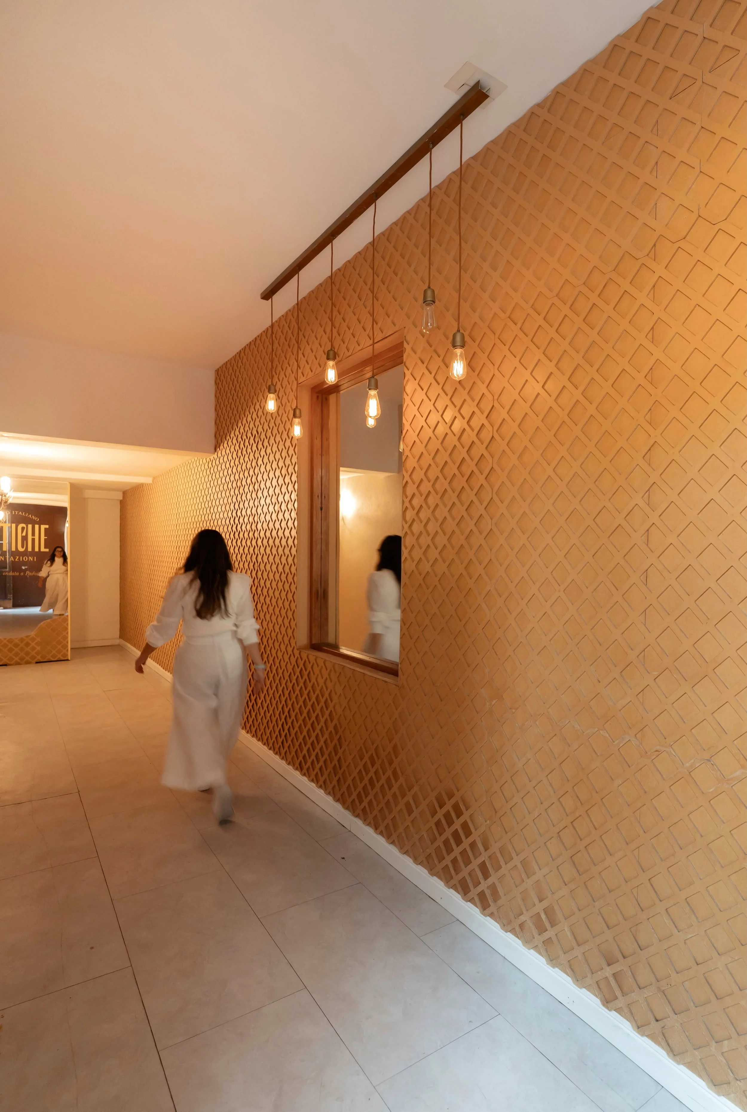 Pasillo con pared de diseño de conchas y luz colgante, mujer caminando con vestido blanco, ventana con reflejo, fondo con cartel y otra mujer caminando.