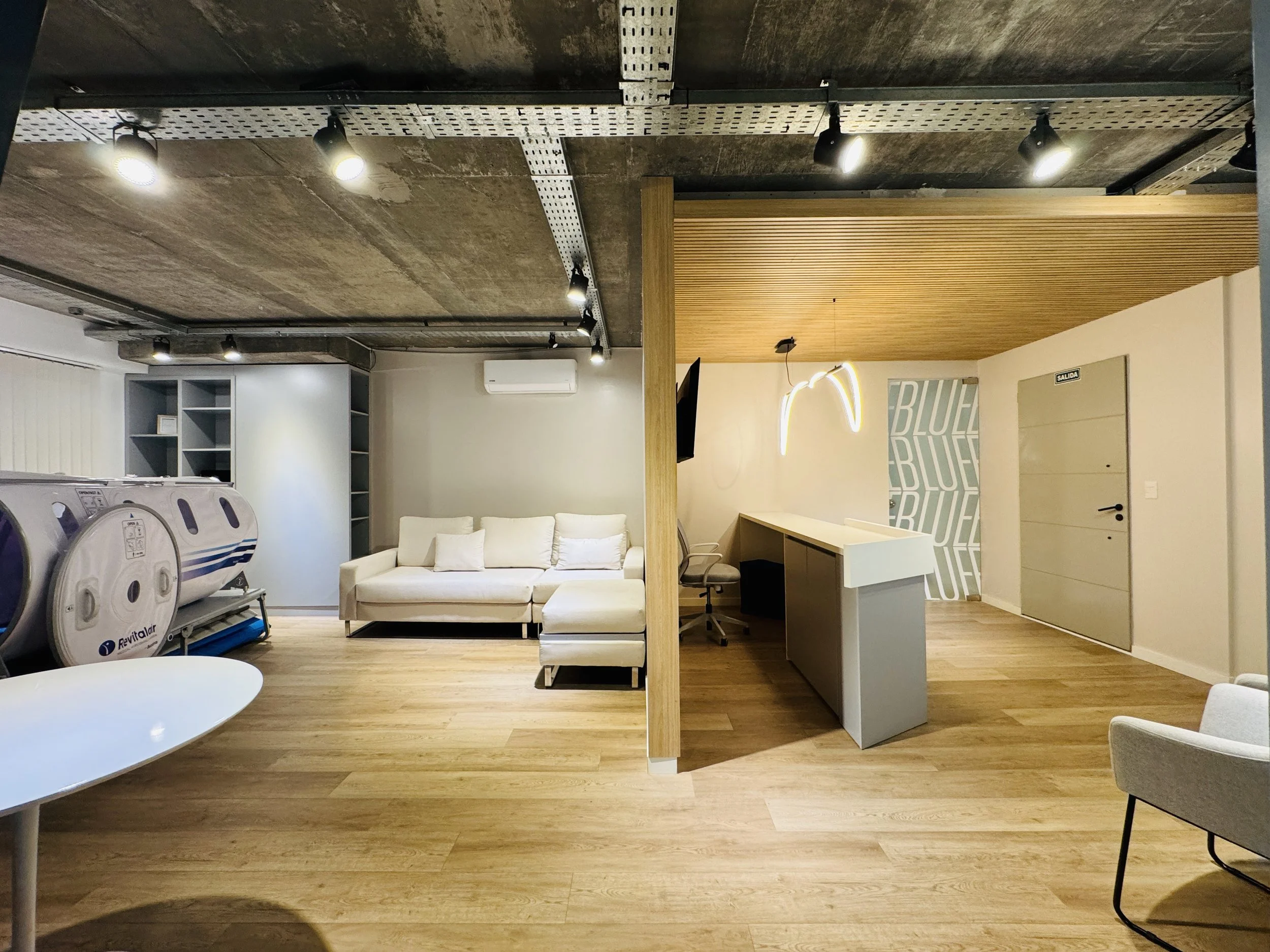 Espacio de oficina moderno con sofá blanco, lavadoras en la esquina izquierda, y área de recepción con escritorio y lámpara