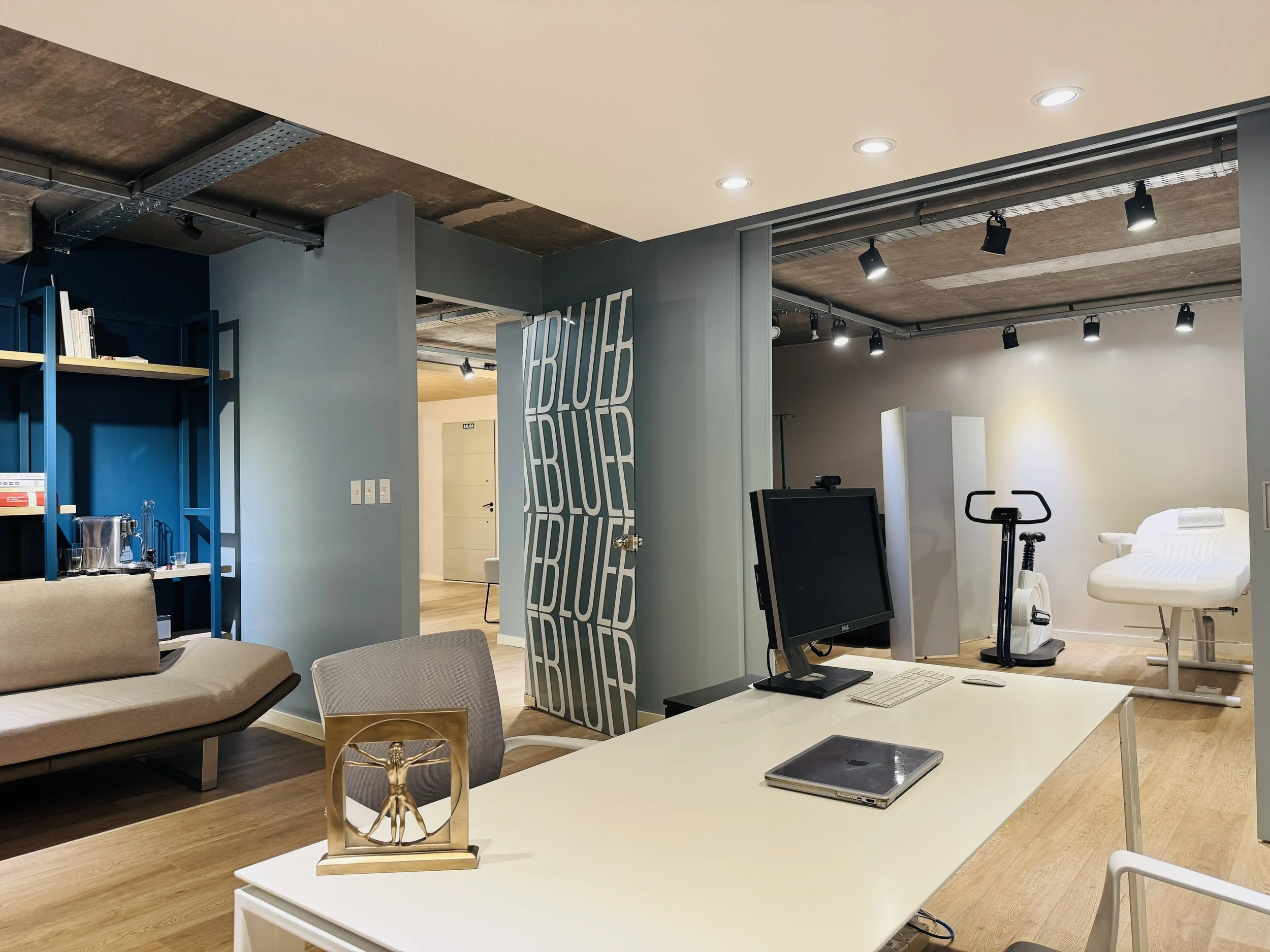 Espacio de oficina con escritorio, monitor, silla, y equipo de ejercicio en una sala de terapia o fisioterapia.