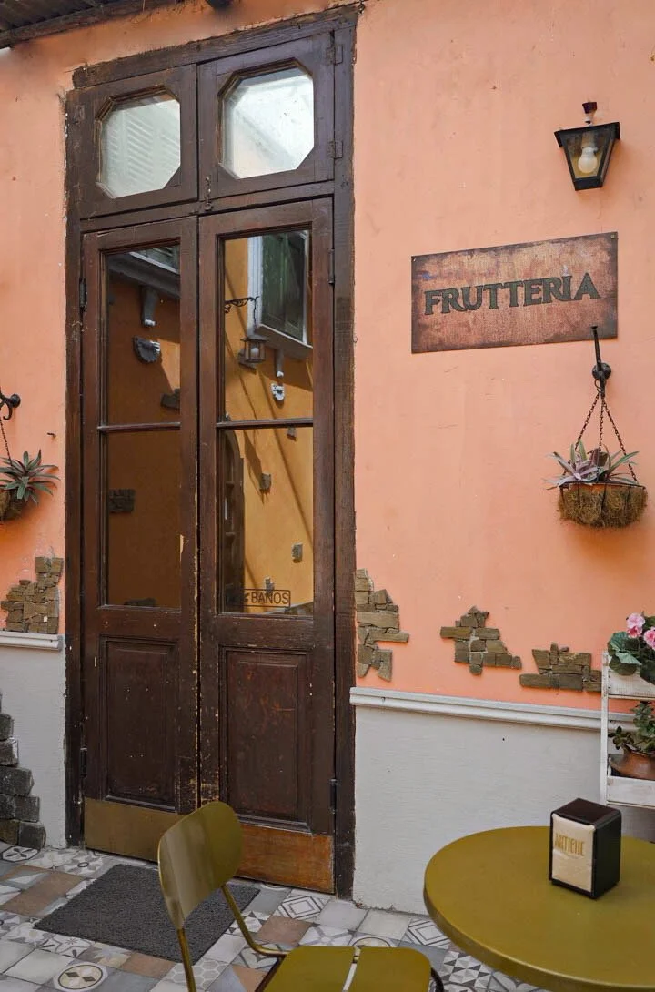 Entrada a una frutería con pared color durazno, cartel de madera que dice 'FRUTTERIA', puerta de madera con ventanas de cristal, plantas en macetas colgantes y en el suelo, y mobiliario de interior.