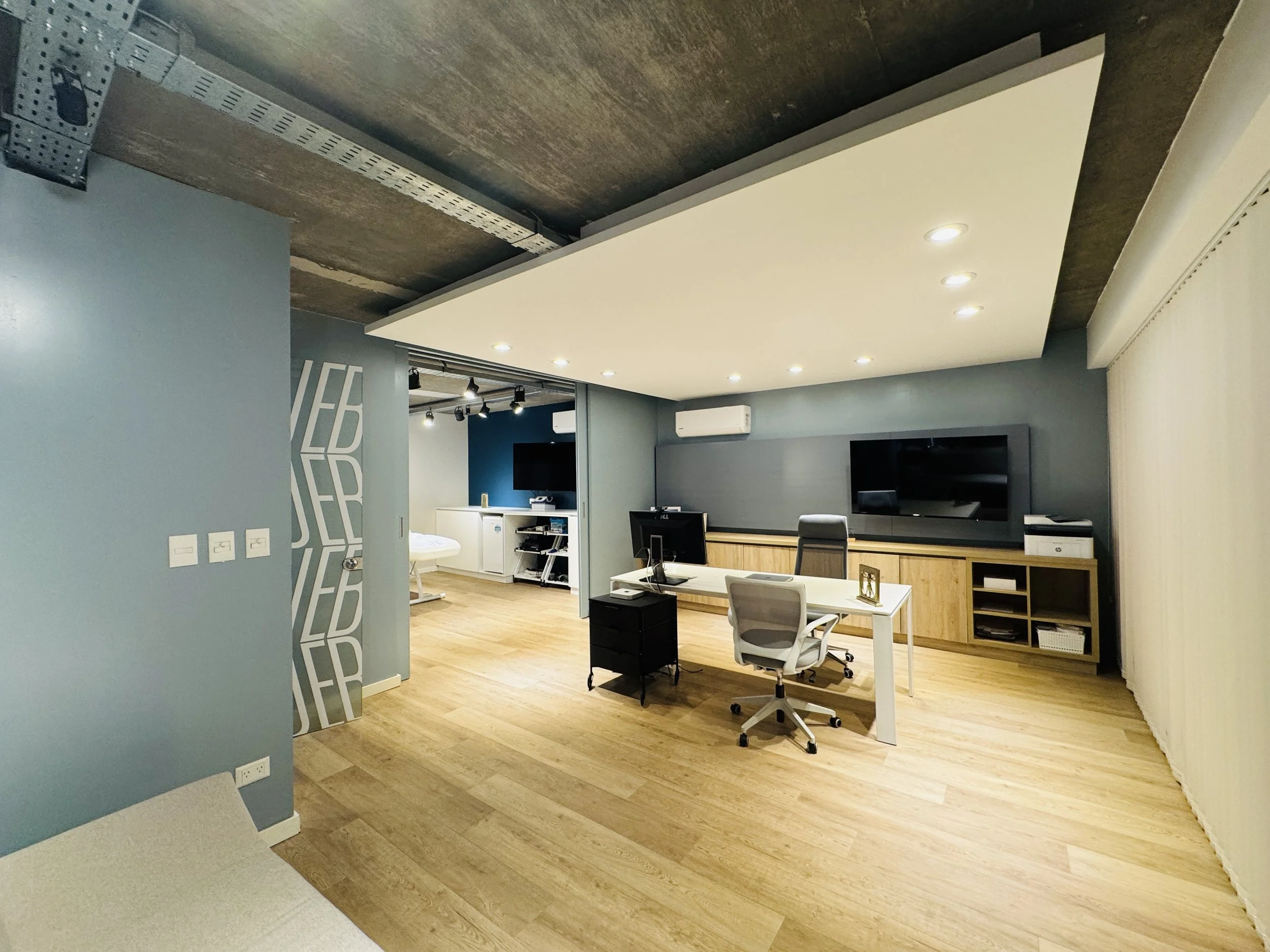 Oficina moderna con escritorio blanco, silla de oficina gris, pantalla grande en la pared, área de trabajo ordenada, suelo de madera clara, y paredes en tonos gris y blanco.