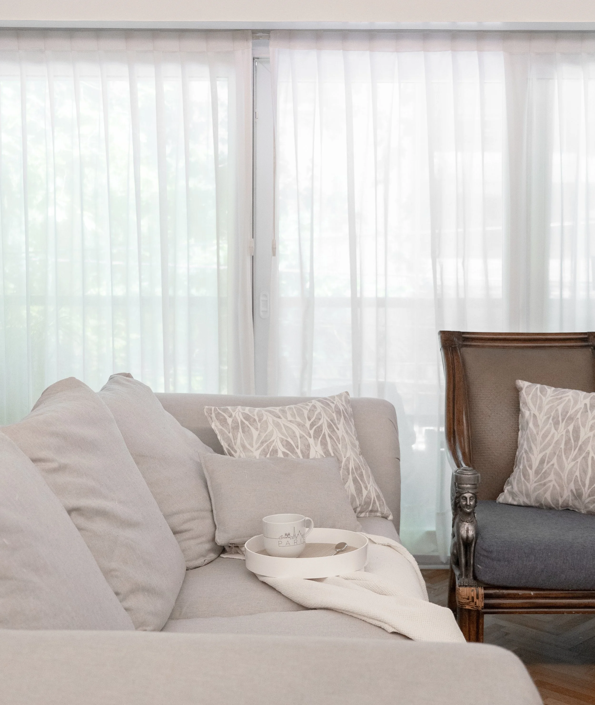 Sala de estar con un sofá gris, cojines decorativos con patrones, una taza en un plato, cortinas blancas en la ventana.