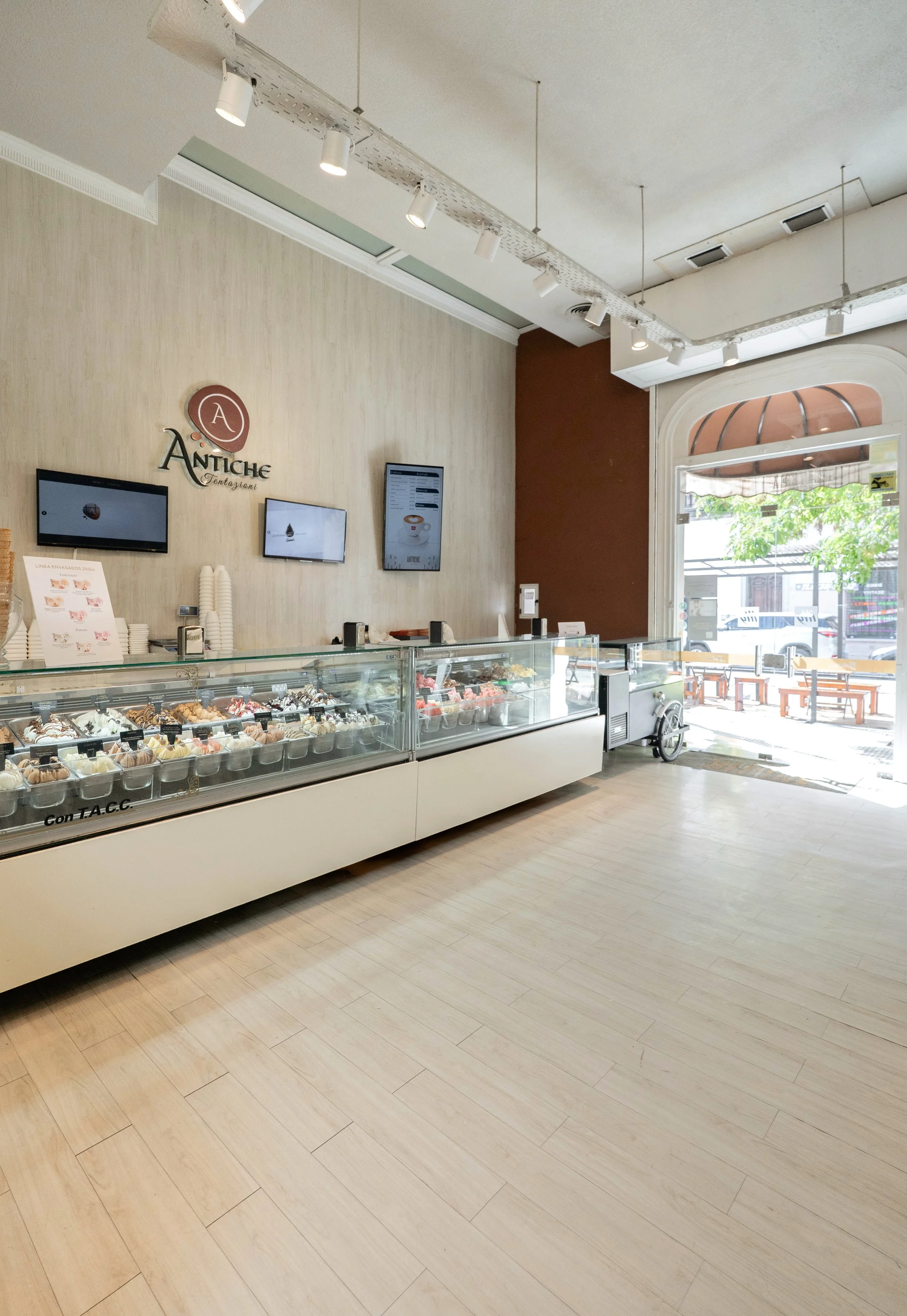 Mostrador de helados en una heladería con envases transparentes llenos de diferentes sabores, en el fondo hay pantallas digitales y una pared con el logotipo Antiche. La entrada de la heladería tiene una puerta de vidrio.