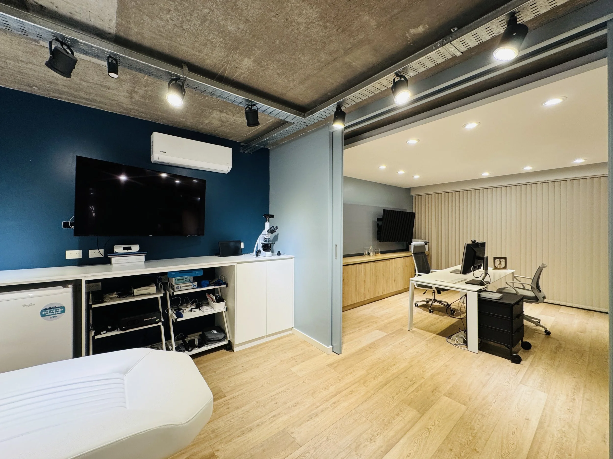 Oficina moderna con paredes azules y blancas, piso de madera clara, con varias mesas con computadoras y sillas ergonómicas, pantalla grande en la pared y cortinas en la ventana.