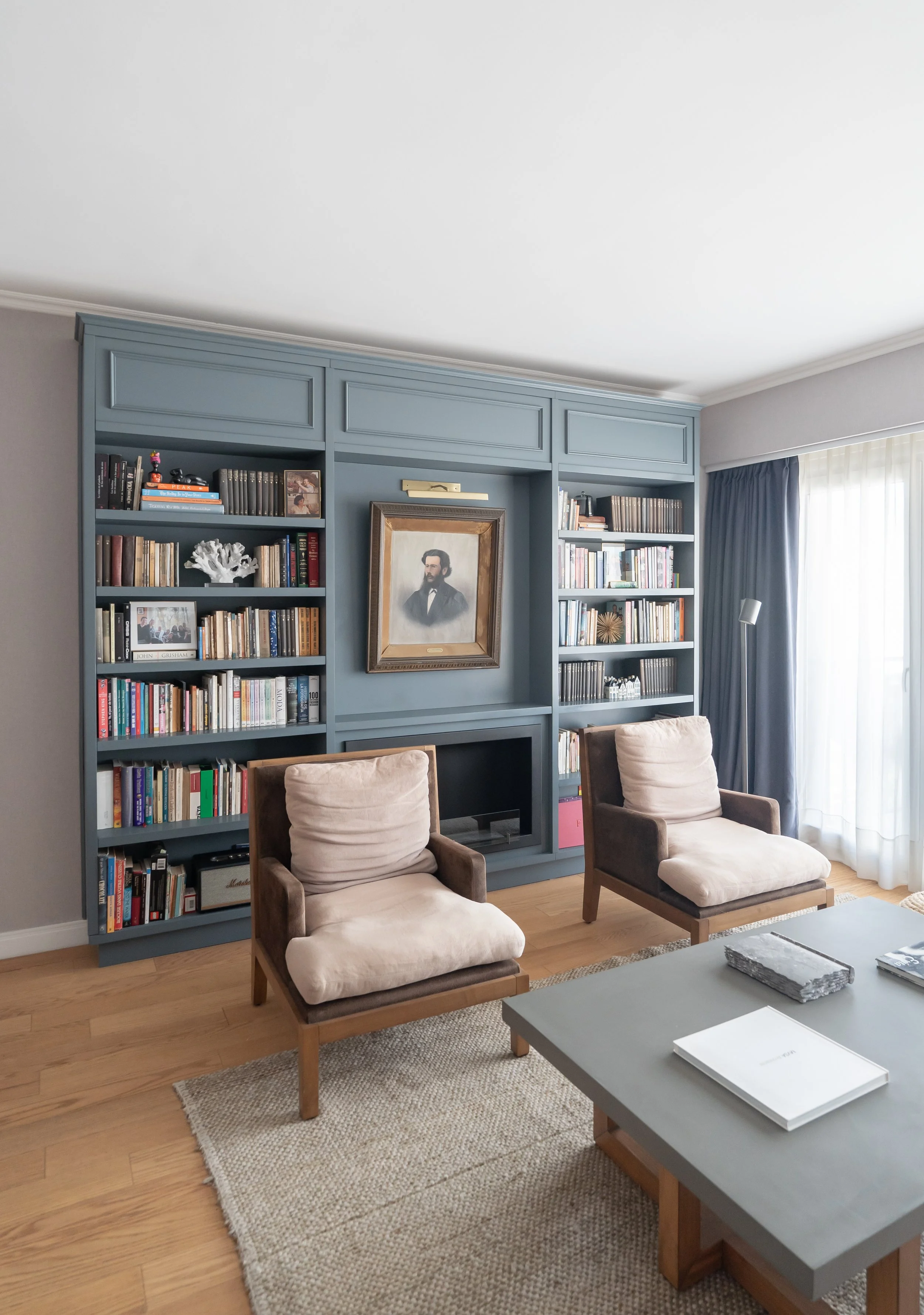 Sala de estar con estantería azul, dos sillones beige, mesa de centro gris y cortinas blancas y azules.