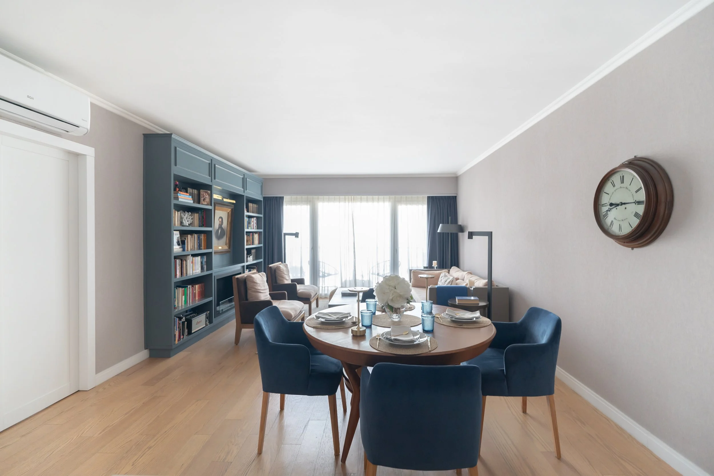 Sala de estar en un apartamento con mesa redonda, seis sillas, un gran librero azul, un reloj de pared y un área de descanso con sillones frente a ventanas con cortinas largas.