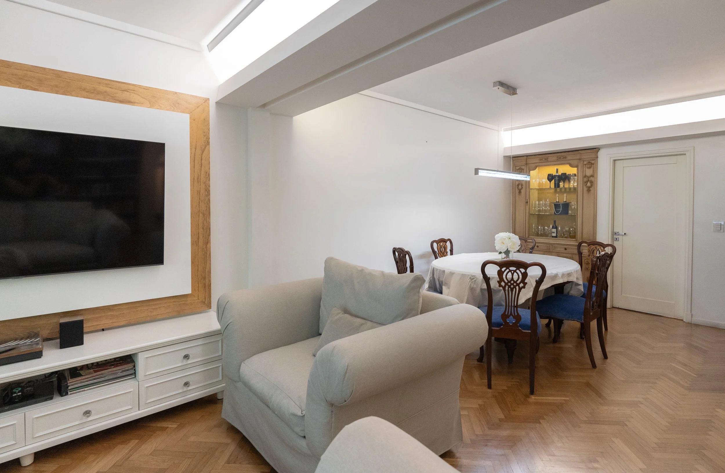 Sala y comedor en una casa con piso de parquet y muebles de madera, un sofá beige, mesa redonda con mantel y flores blancas, televisor en la pared, y una vitrina de madera con botellas y cristalería.