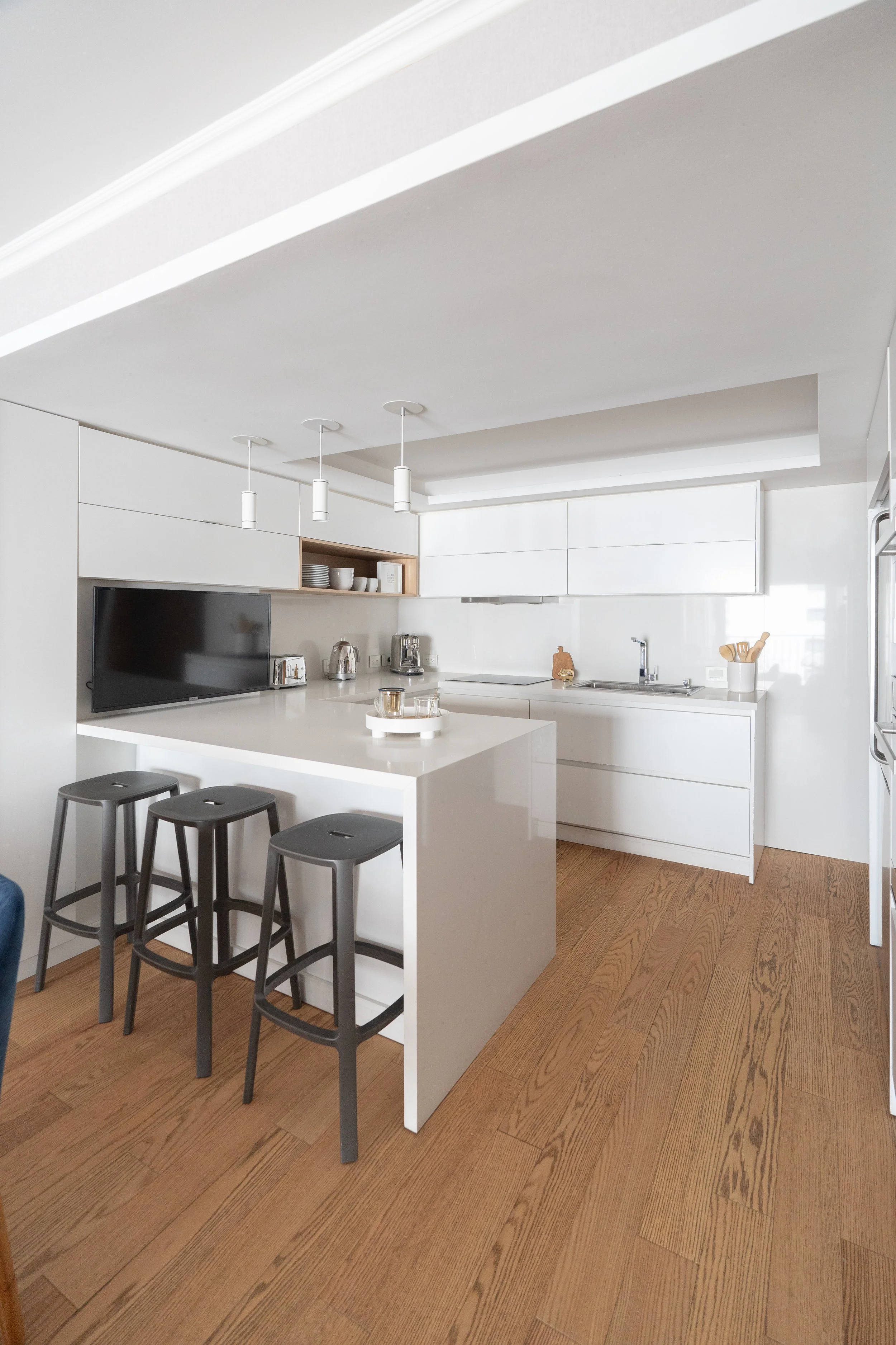 Cocina moderna y minimalista con encimera blanca, armarios blancos, piso de madera, una televisión, y utensilios de cocina.