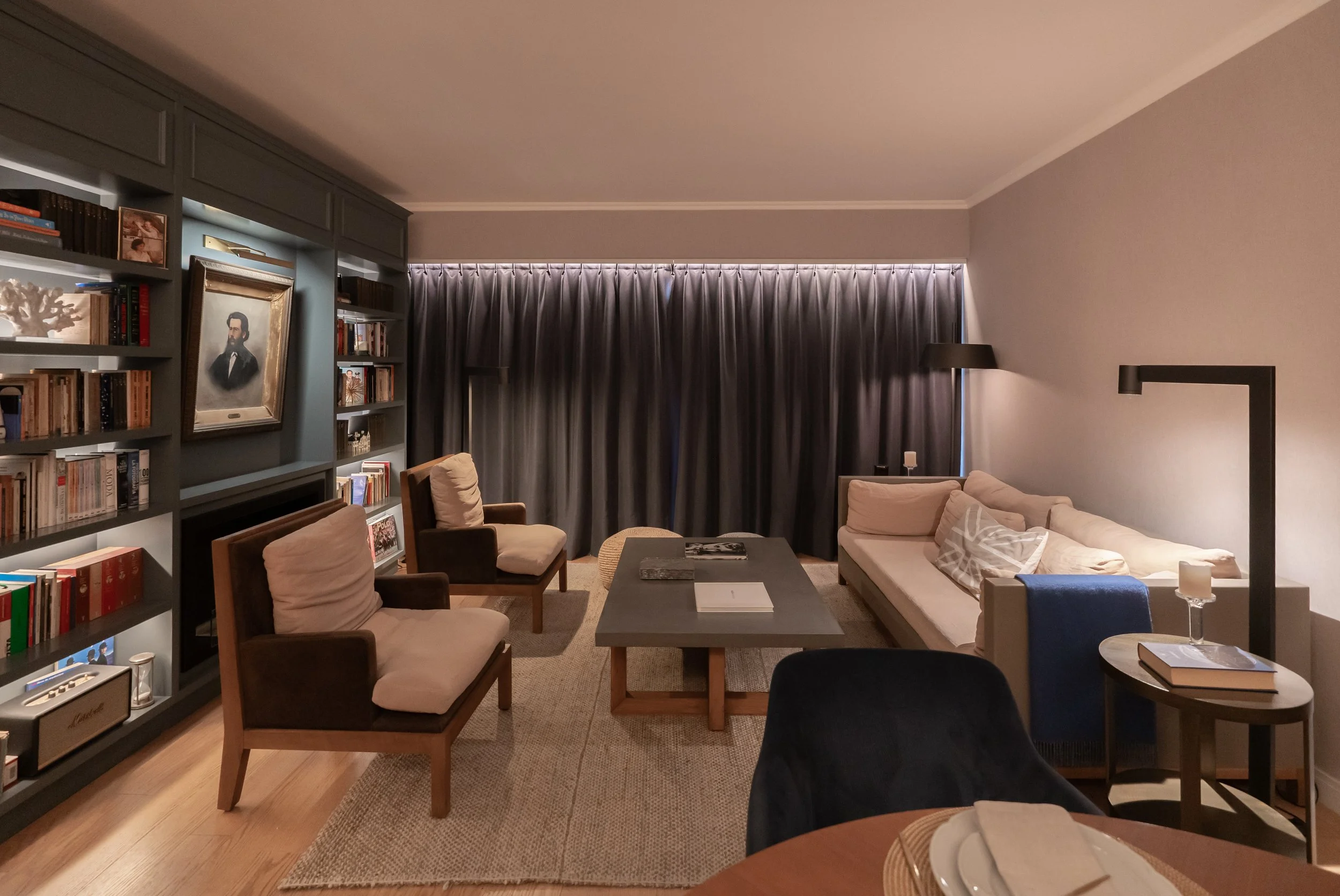 Sala de estar con sillones, una mesa de café, estantería con libros, cortinas oscuras y una esquina con lámpara de piso.