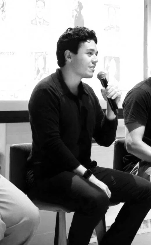 Mardoqueo Arteaga sitting on a chair, holding a microphone and speaking at an event or panel, with a presentation slide in the background.