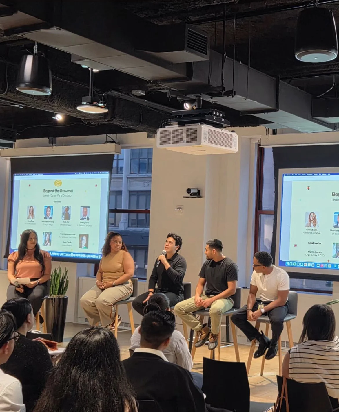 Panel discussion with five people on stage, two large screens displaying presentation slides, audience seated facing the stage in an indoor venue with large windows.