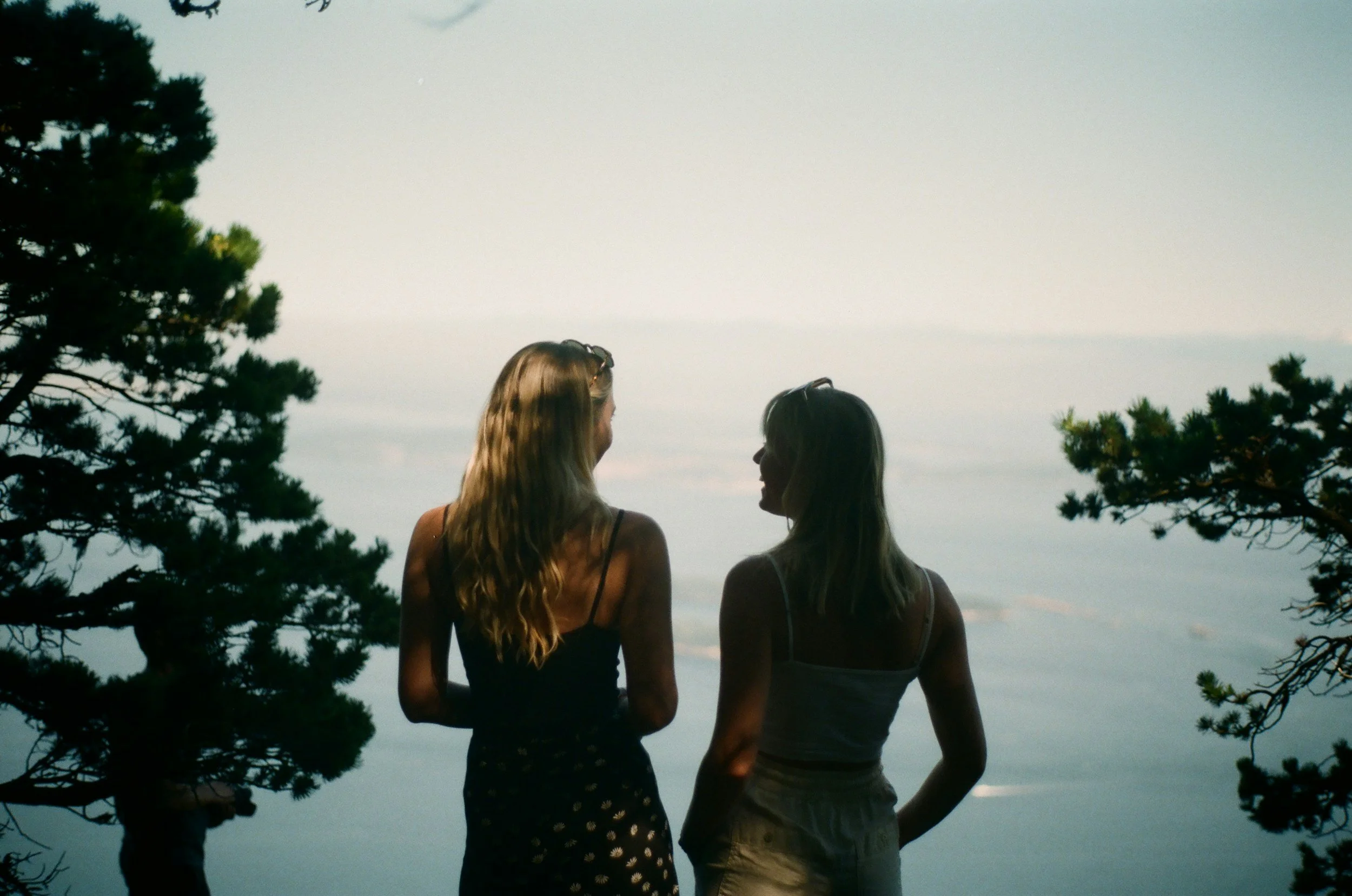 Two women with long hair, wearing tank tops, standing near trees and looking at each other with a body of water in the background.