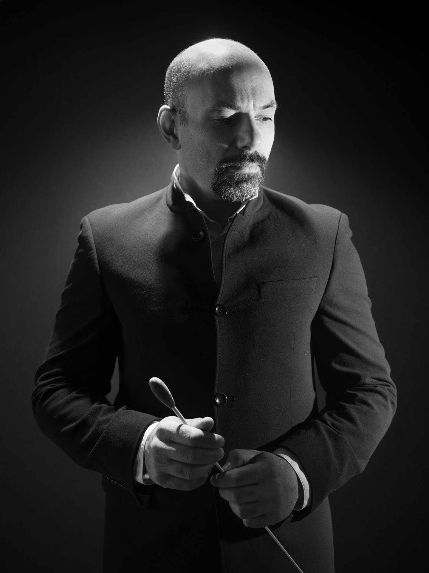 Black and white portrait of a man with a goatee and bald head, wearing a dark suit, holding a conductor's baton, looking down contemplatively.
