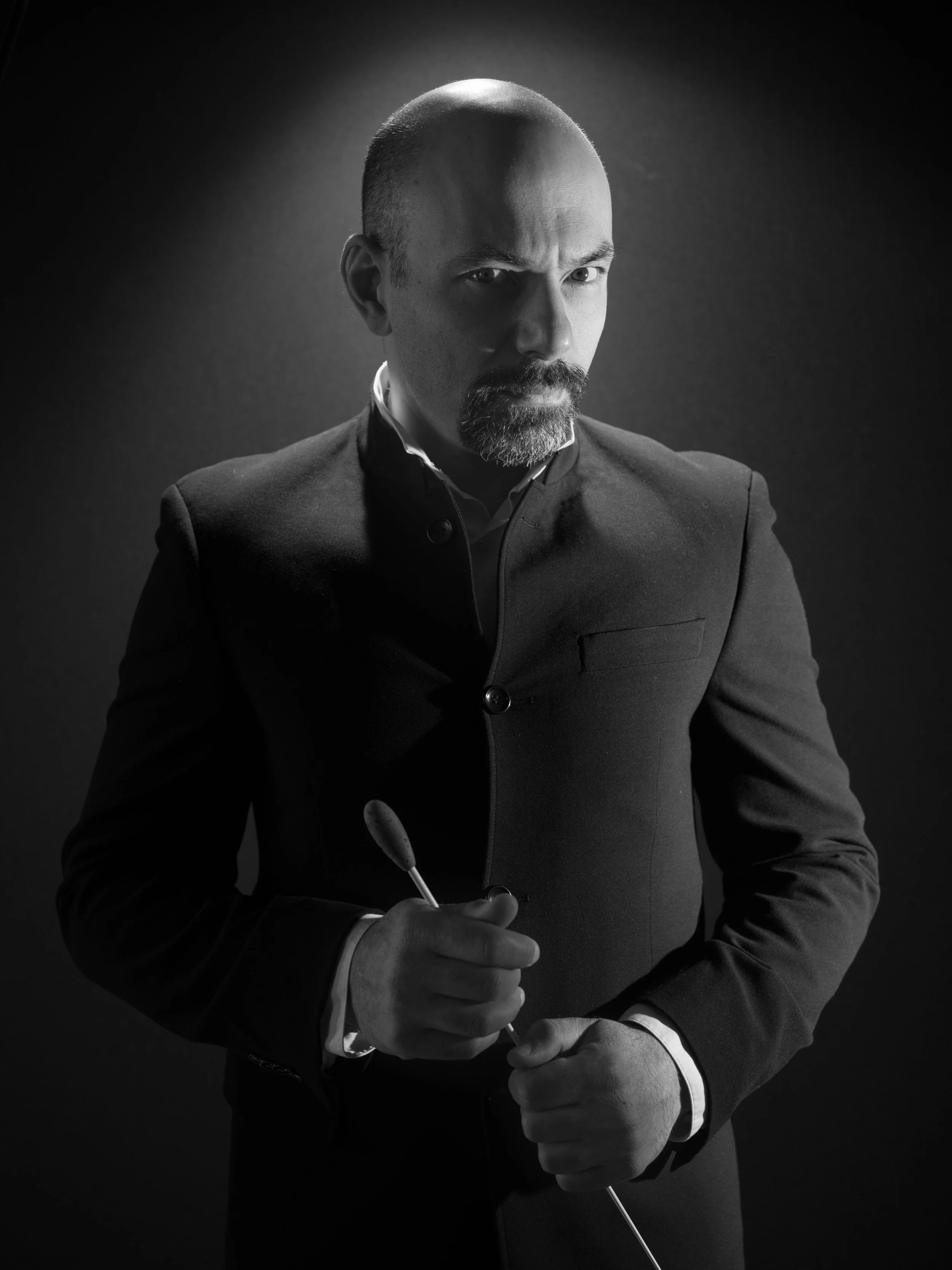 A black and white portrait of a serious man with a bald head and a beard, wearing a formal suit, holding a dart, in a dramatic lighting setting.