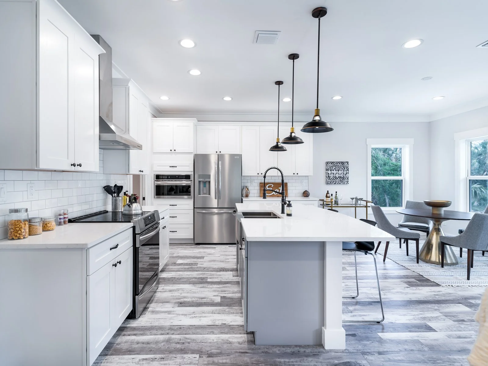 Bright modern kitchen with white cabinets, stainless steel oven and refrigerator, island with white countertop, black pendant lights, and dining area with gray chairs and round table near large windows.