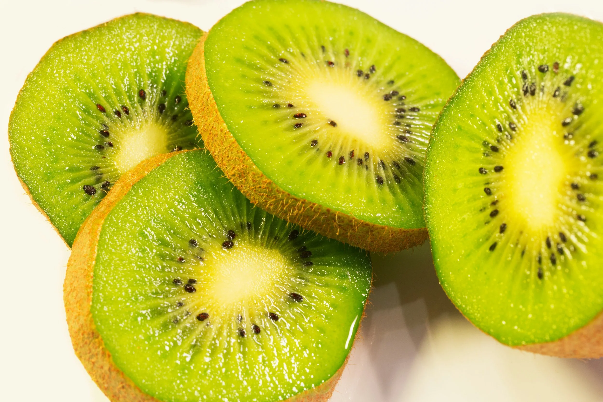 Multiple slices of kiwi fruit with bright green flesh, small black seeds, and brown fuzzy skin, arranged overlapping on a white background.